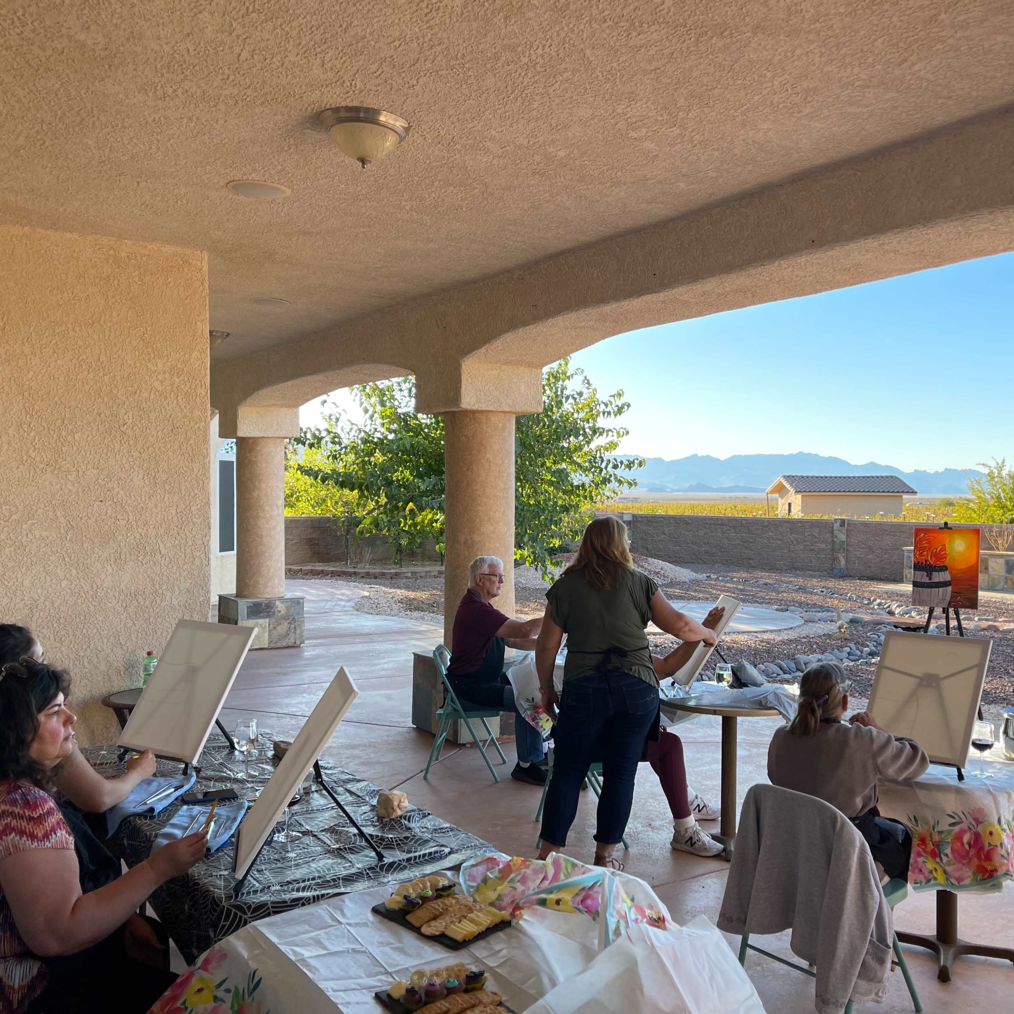A group of people participates in a painting class outdoors under a covered patio with a mountain view.