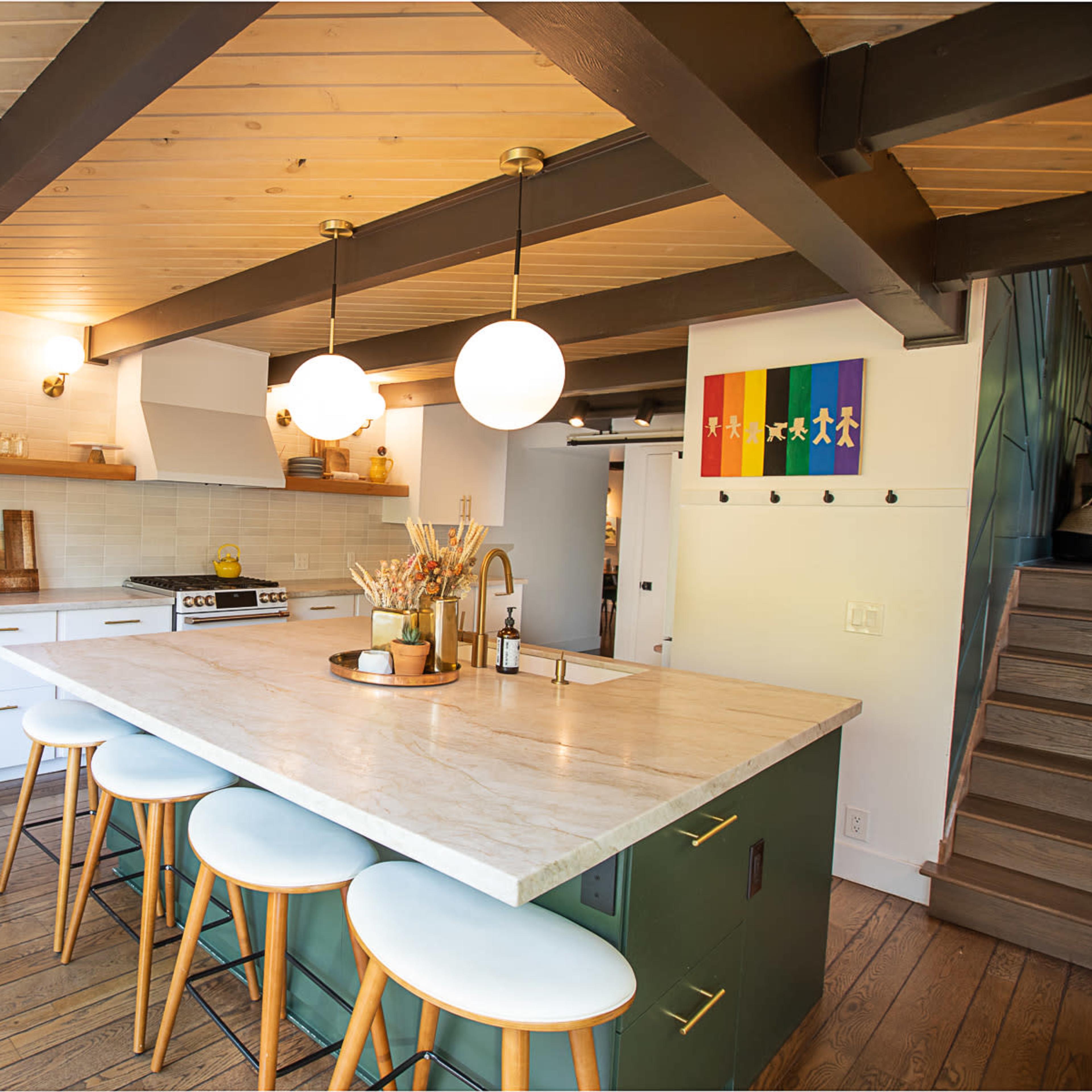 A modern kitchen features a central island with seating, white cabinetry, wooden beams, and a colorful wall decoration.