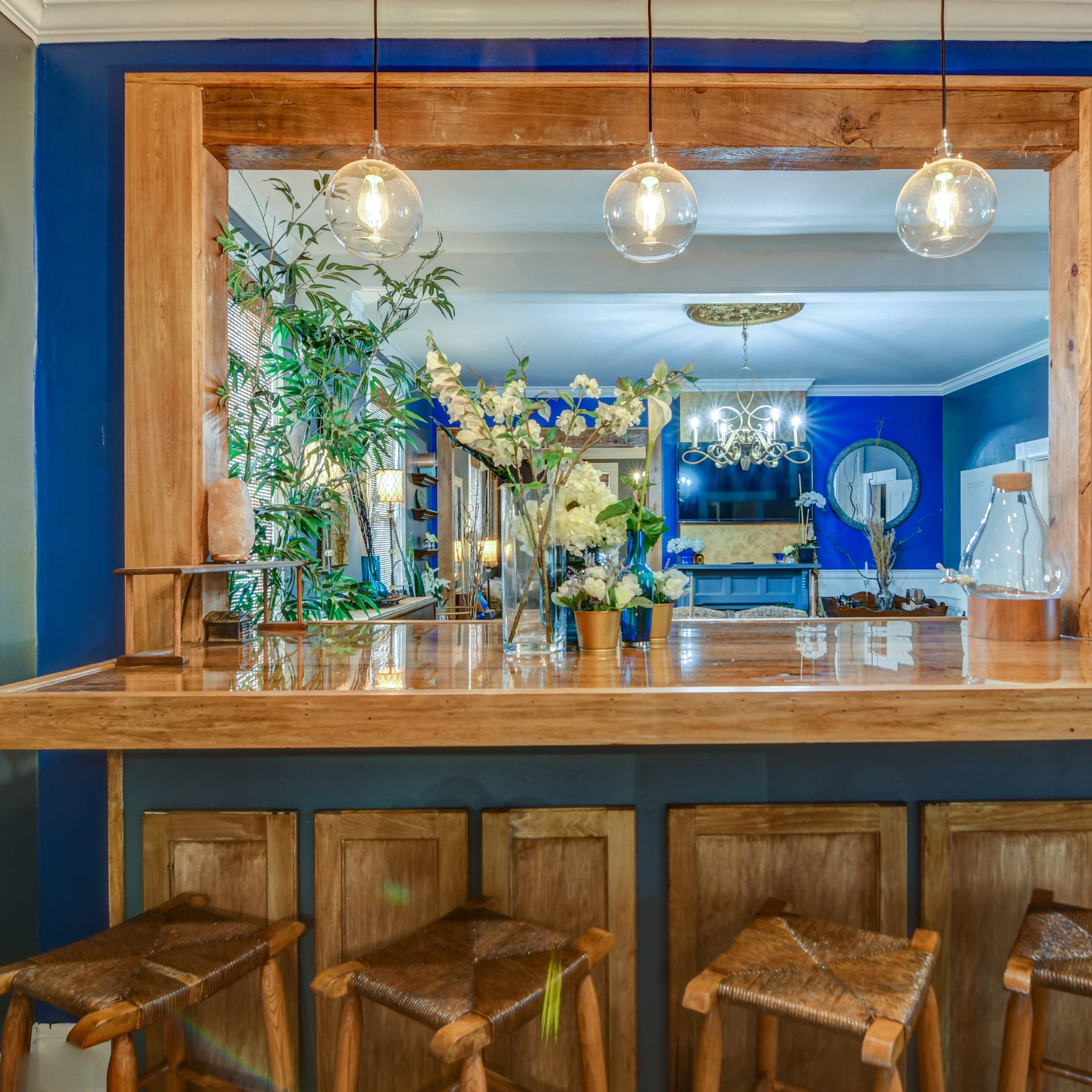 A wooden bar with three stools is lit by pendant lights and features a vase of flowers, set against a blue wall with plants in the background.