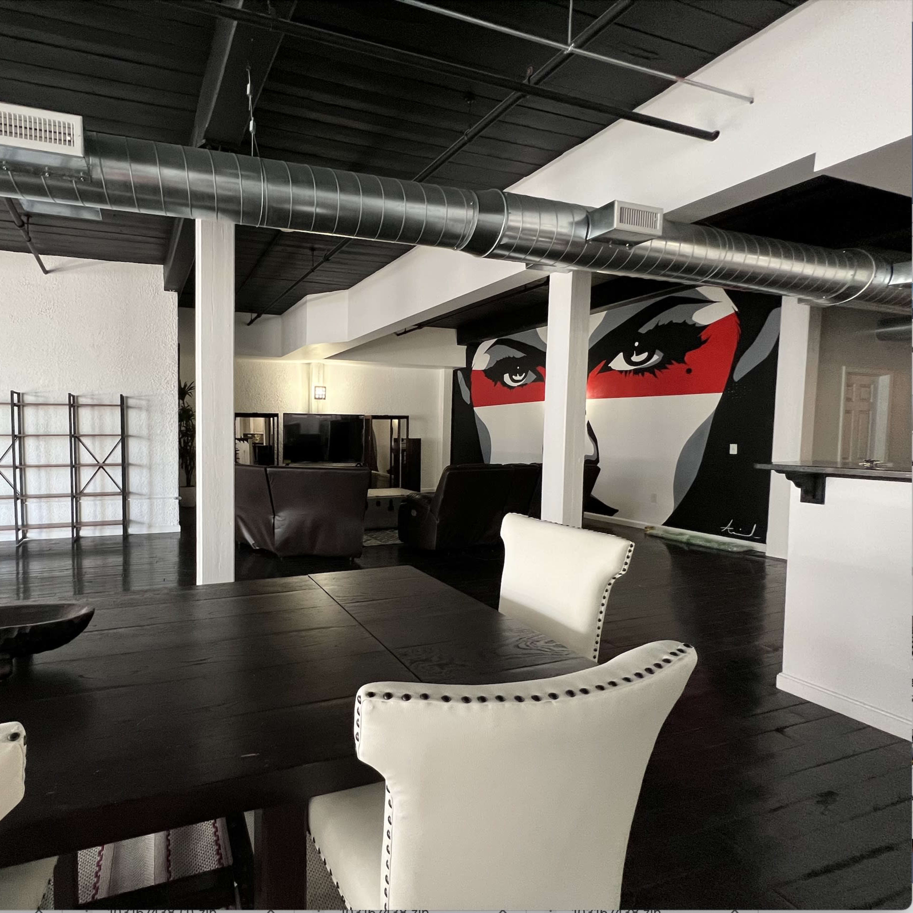 A spacious interior with a dining table and chairs in the foreground, a large wall mural of a face in the background, and various furniture pieces in an open living area.