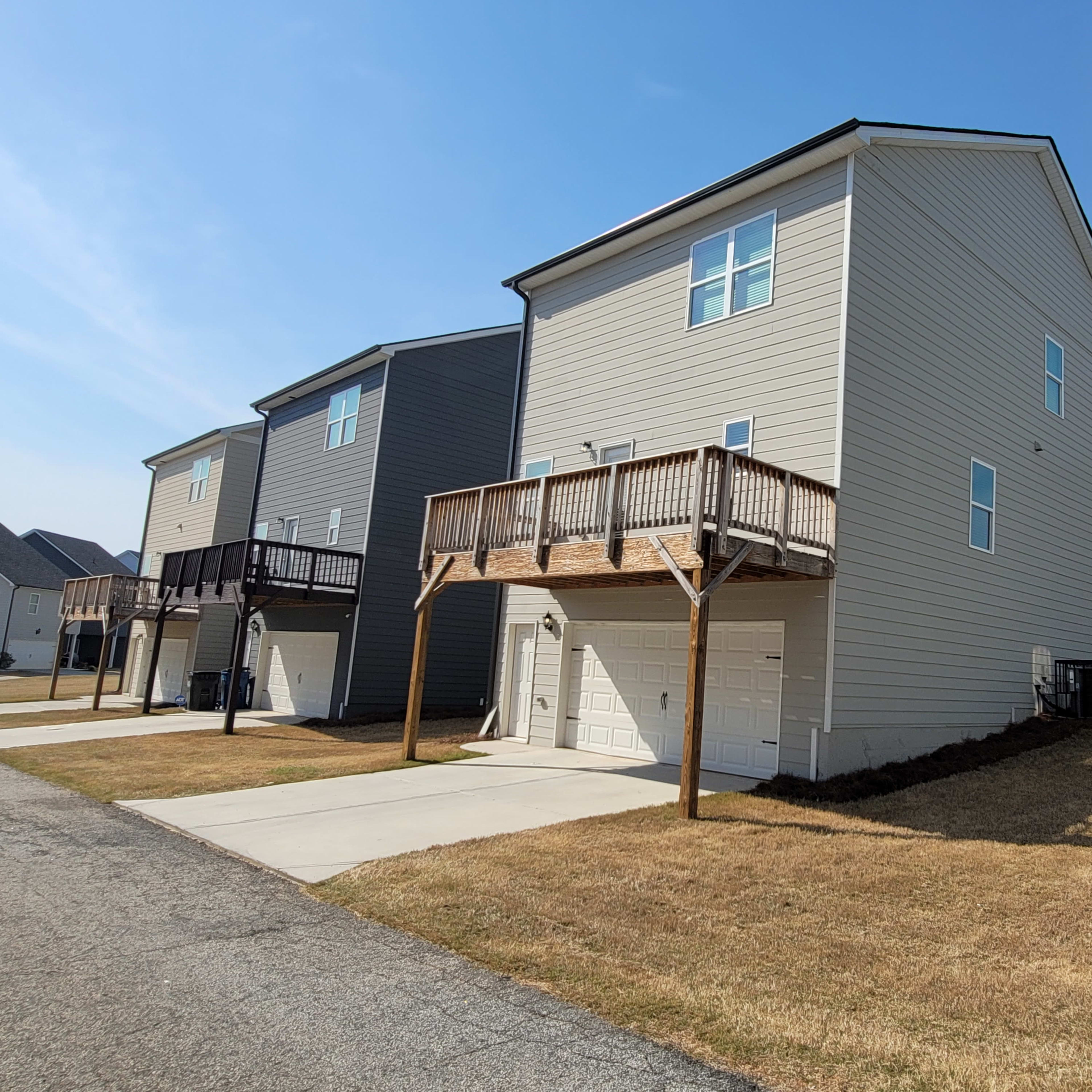 A two-story house with a wooden deck is situated next to a gravel road in a suburban neighborhood.