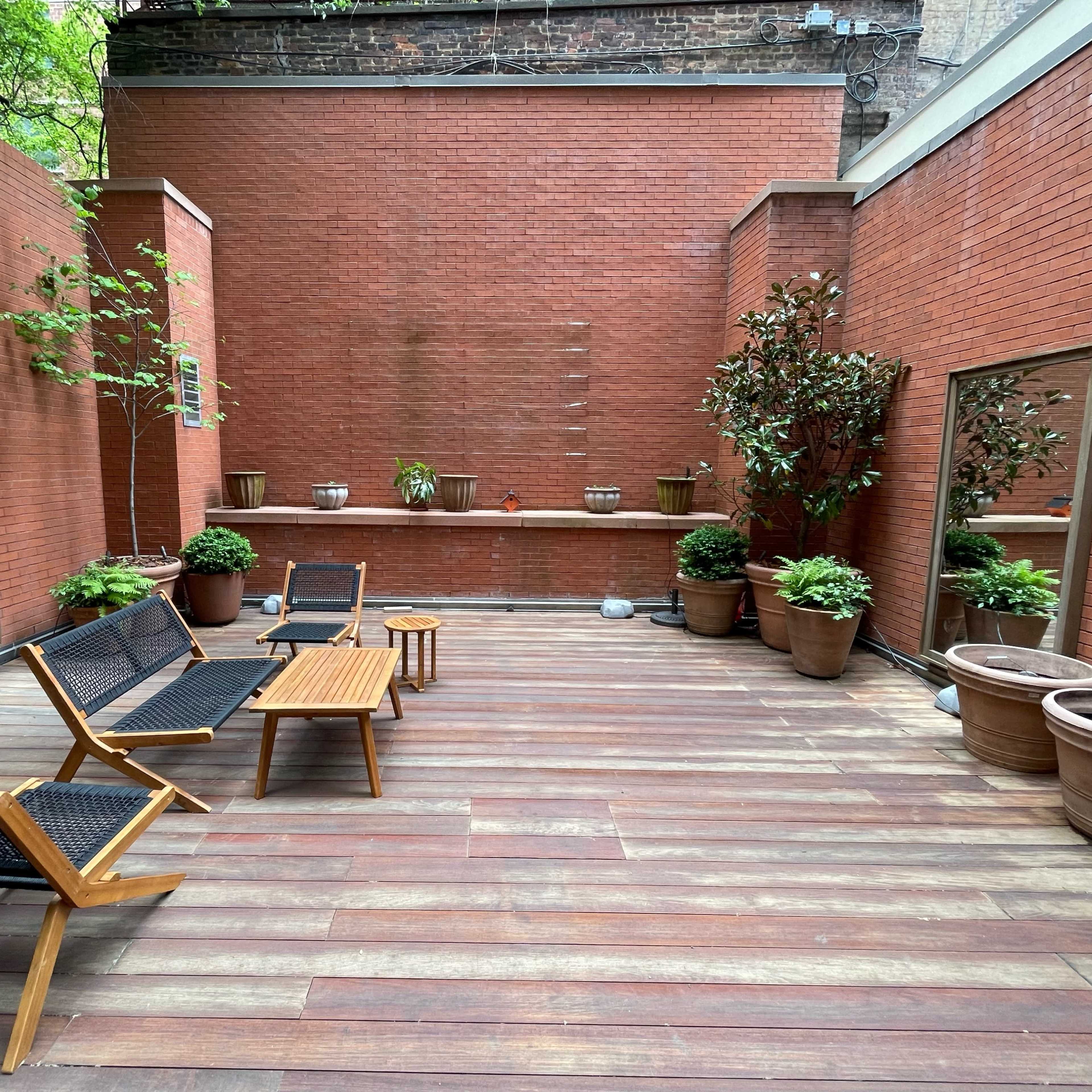 A small outdoor patio with wooden flooring, potted plants, and two seating areas flanked by mirrors on the brick walls.