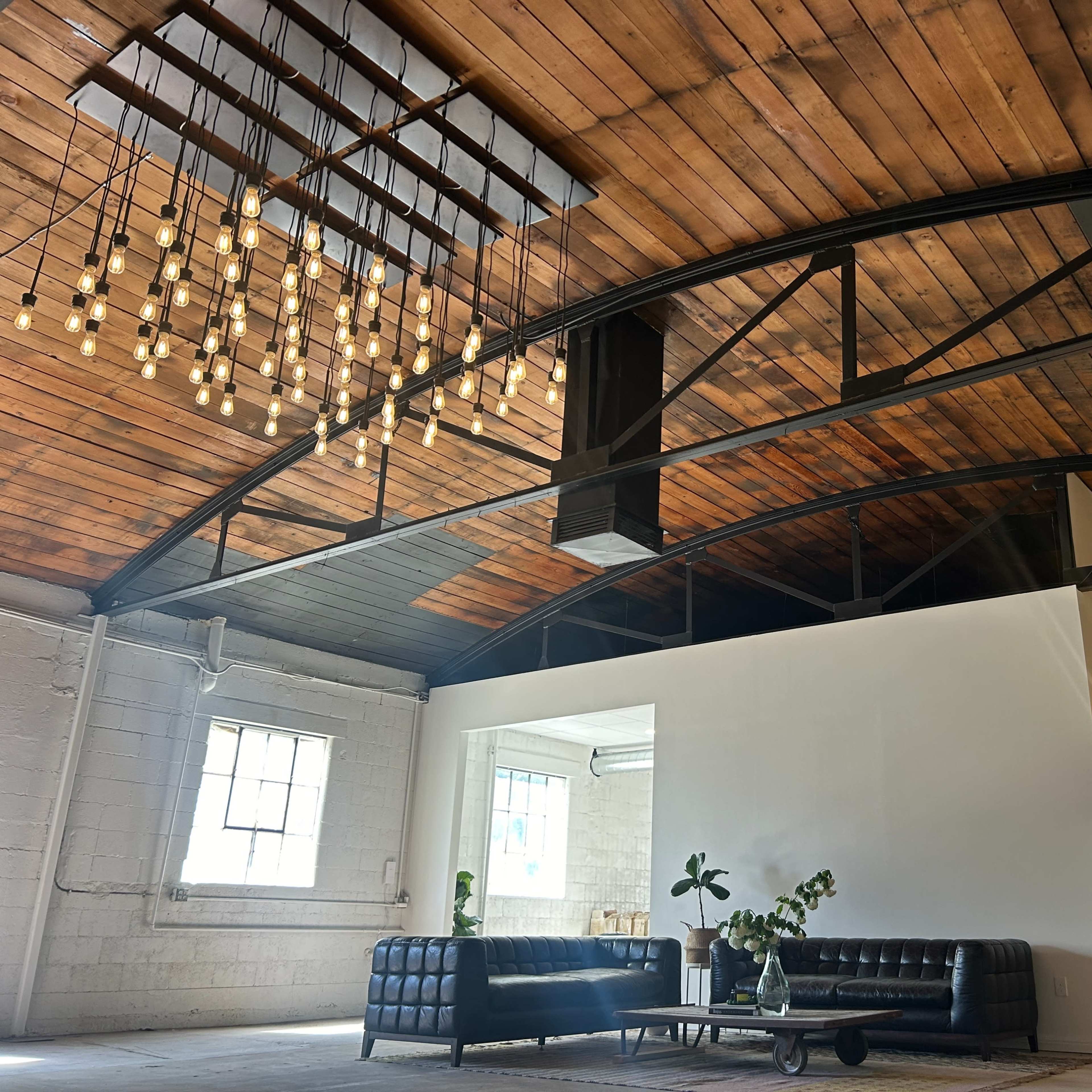 An industrial-style interior featuring a high wooden ceiling with pendant lights, a black leather sofa, and a large white wall in an open space.