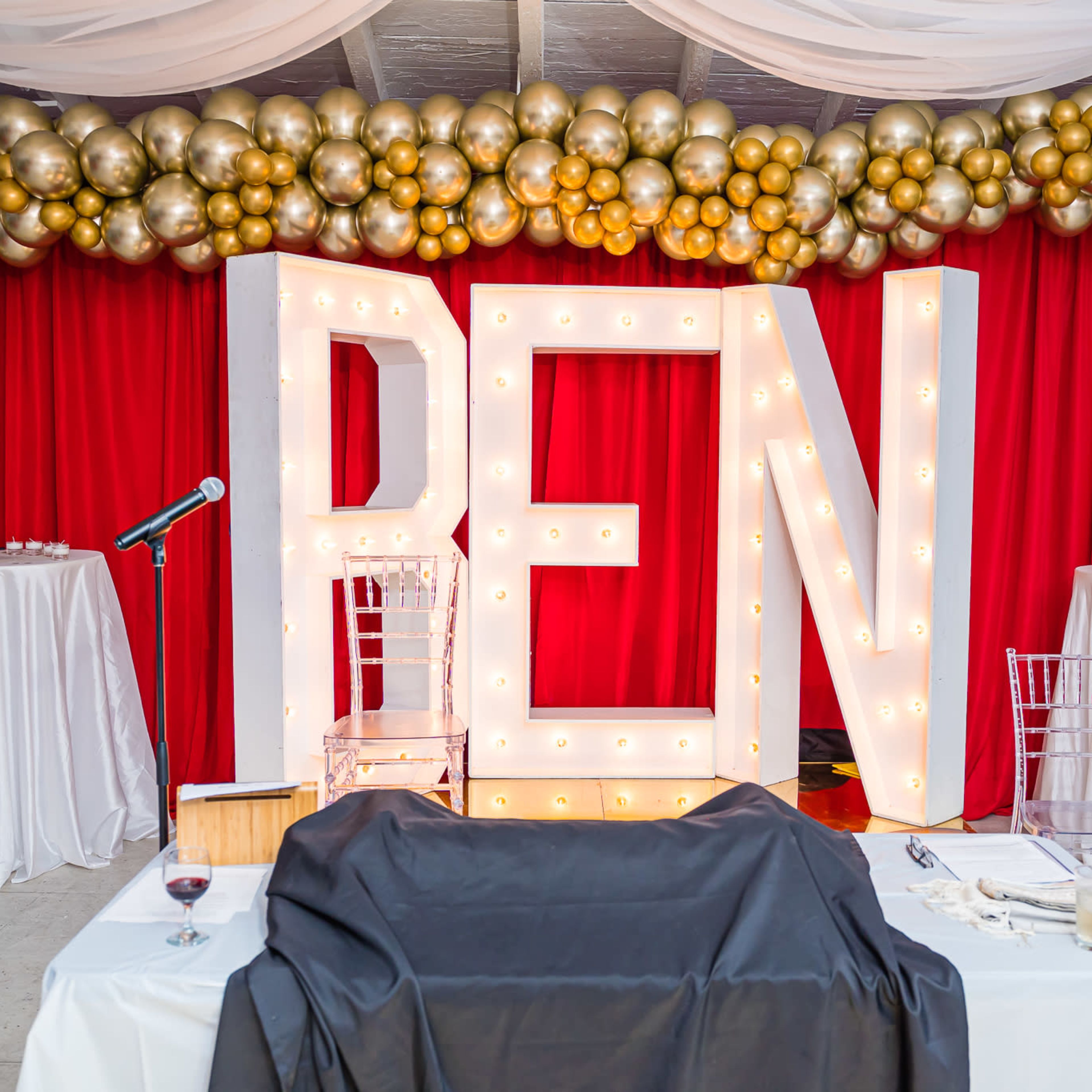 The scene features large illuminated letters spelling "REN" against a backdrop of red curtains and a decorative arrangement of gold and silver balloons.