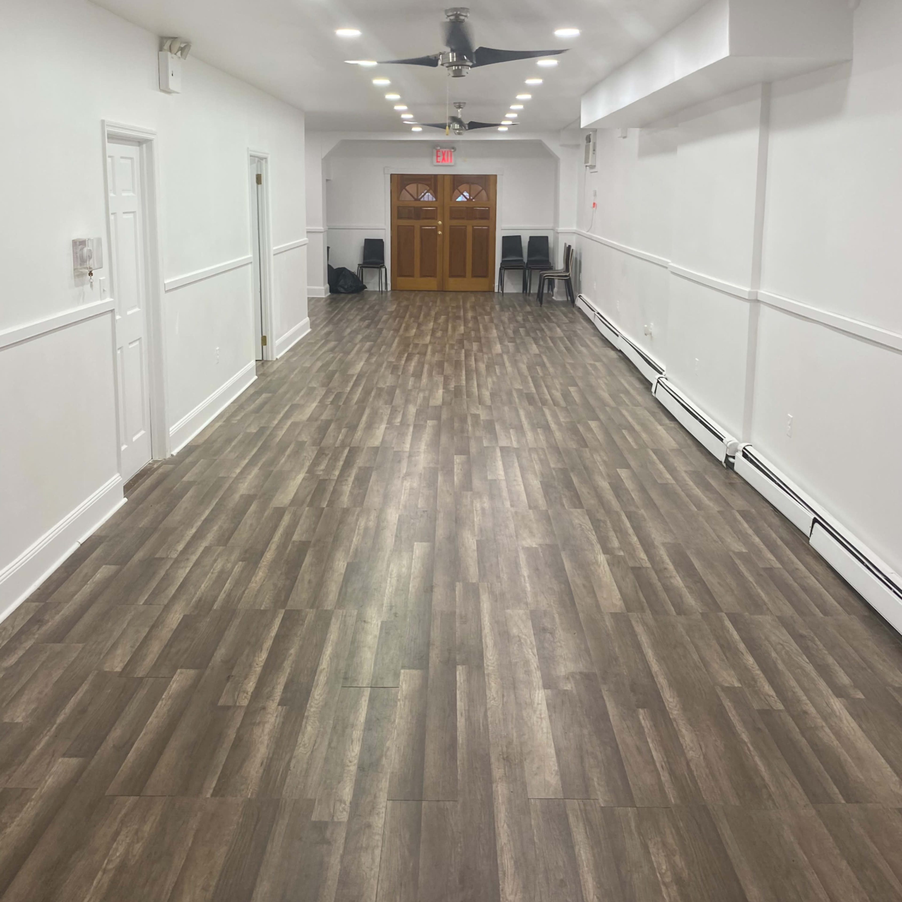 A long, empty room with wooden flooring, white walls, and a door at the far end leading outside.