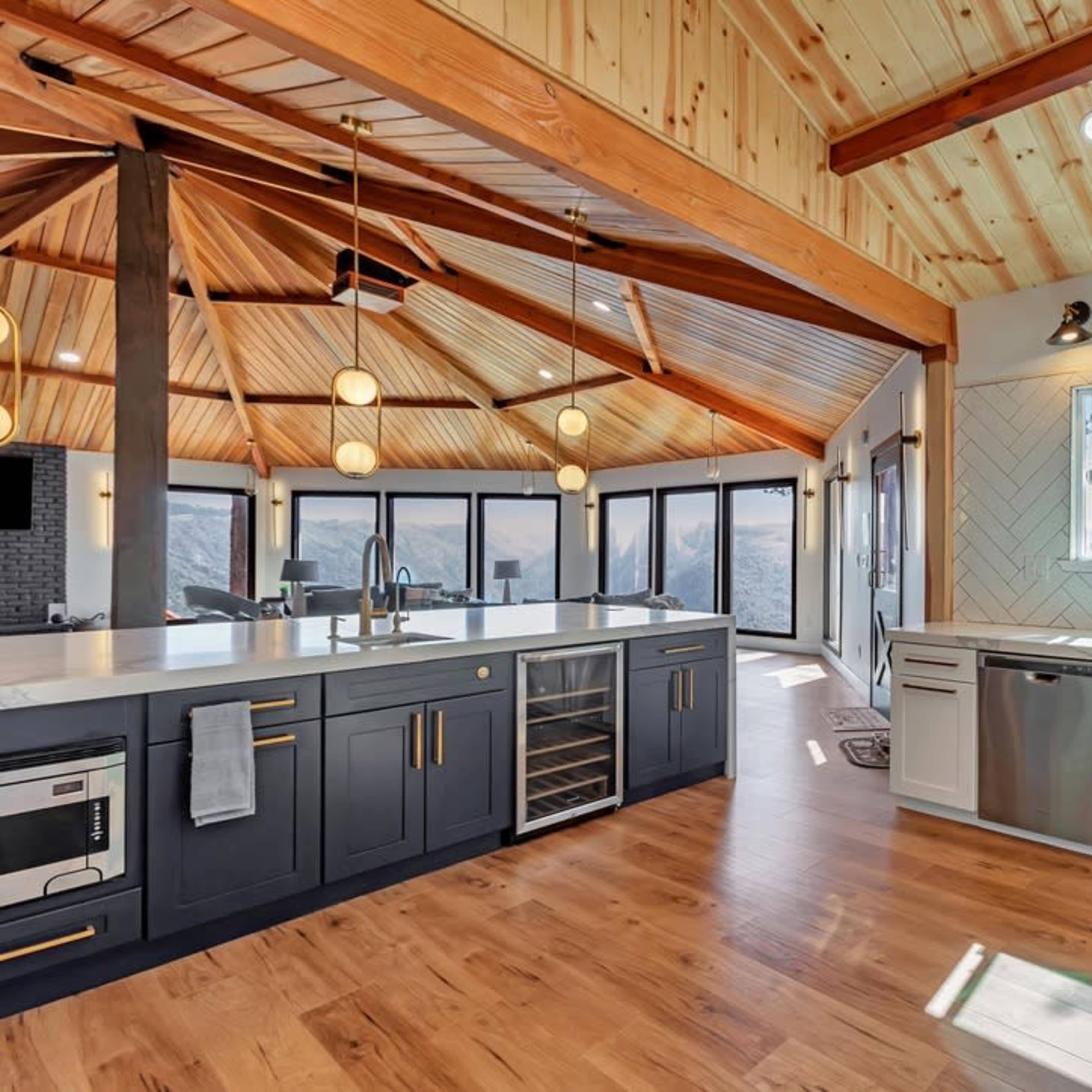 A modern kitchen with a dark blue island, white cabinetry, and large windows offering a view of the outdoors, all highlighted by a wood-paneled ceiling.