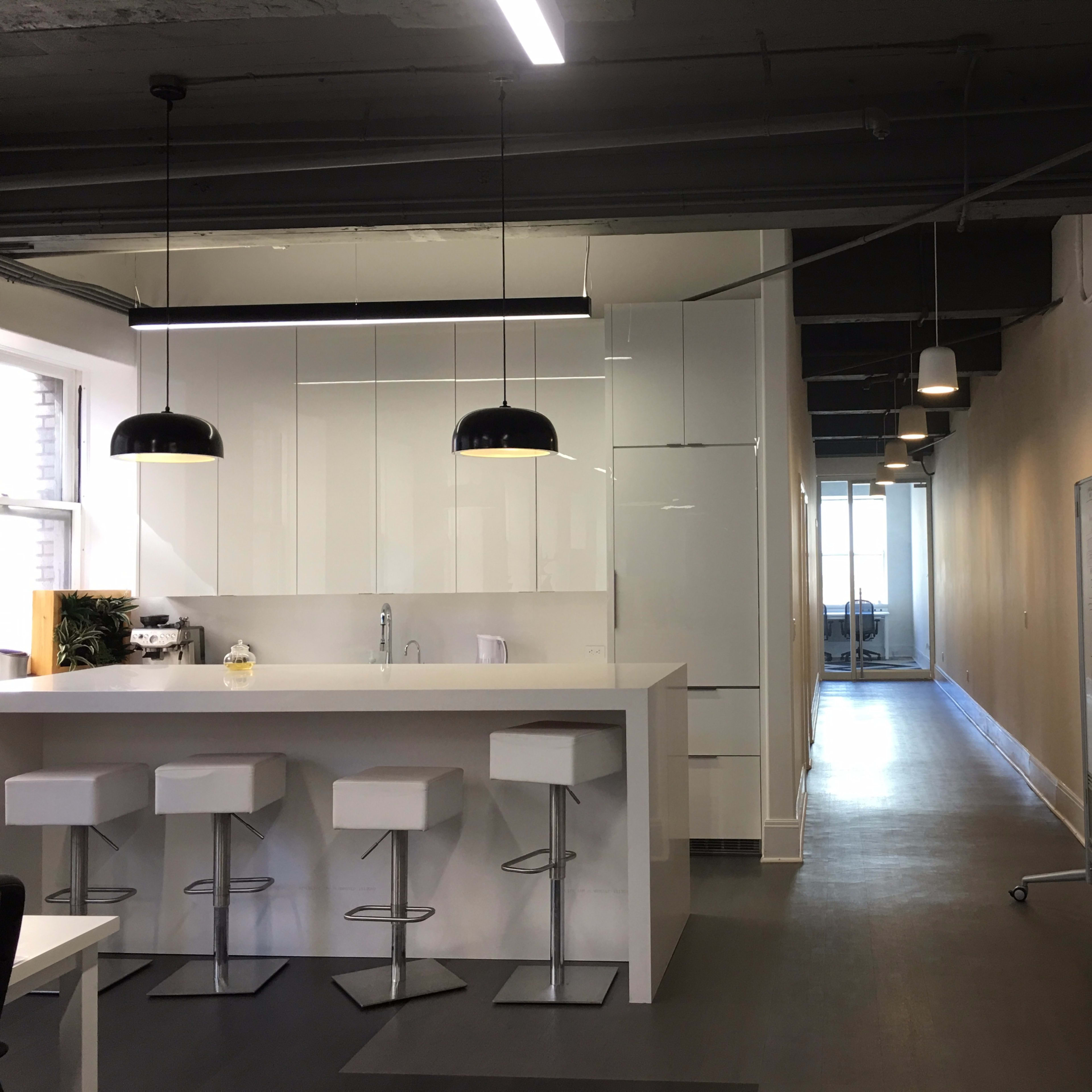 A modern office space featuring a white kitchen island with bar stools, bright lighting, and a corridor leading to a large window at the far end.