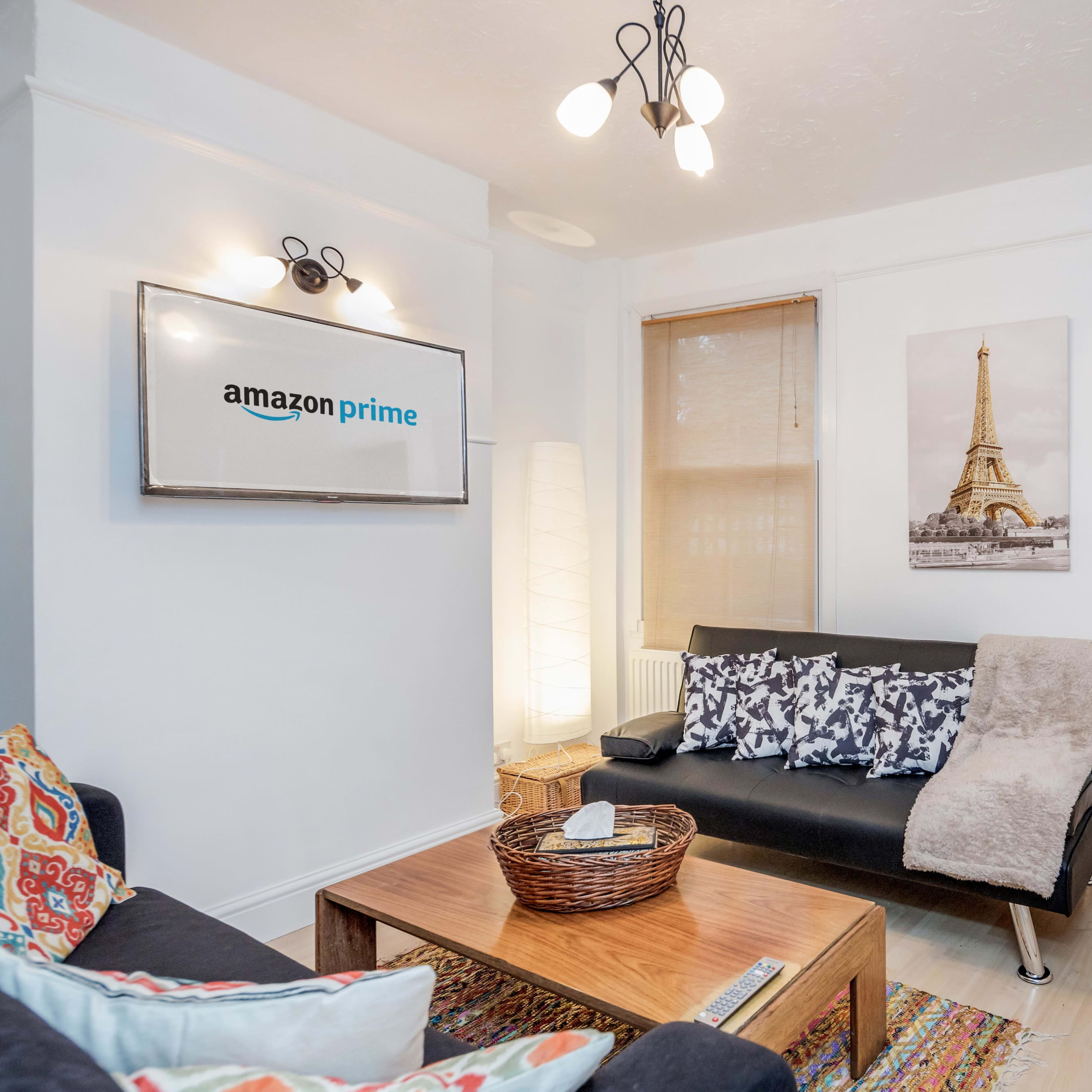 A living room with a black couch, a coffee table, an Amazon Prime sign on the wall, and a doorway leading to a kitchen area.