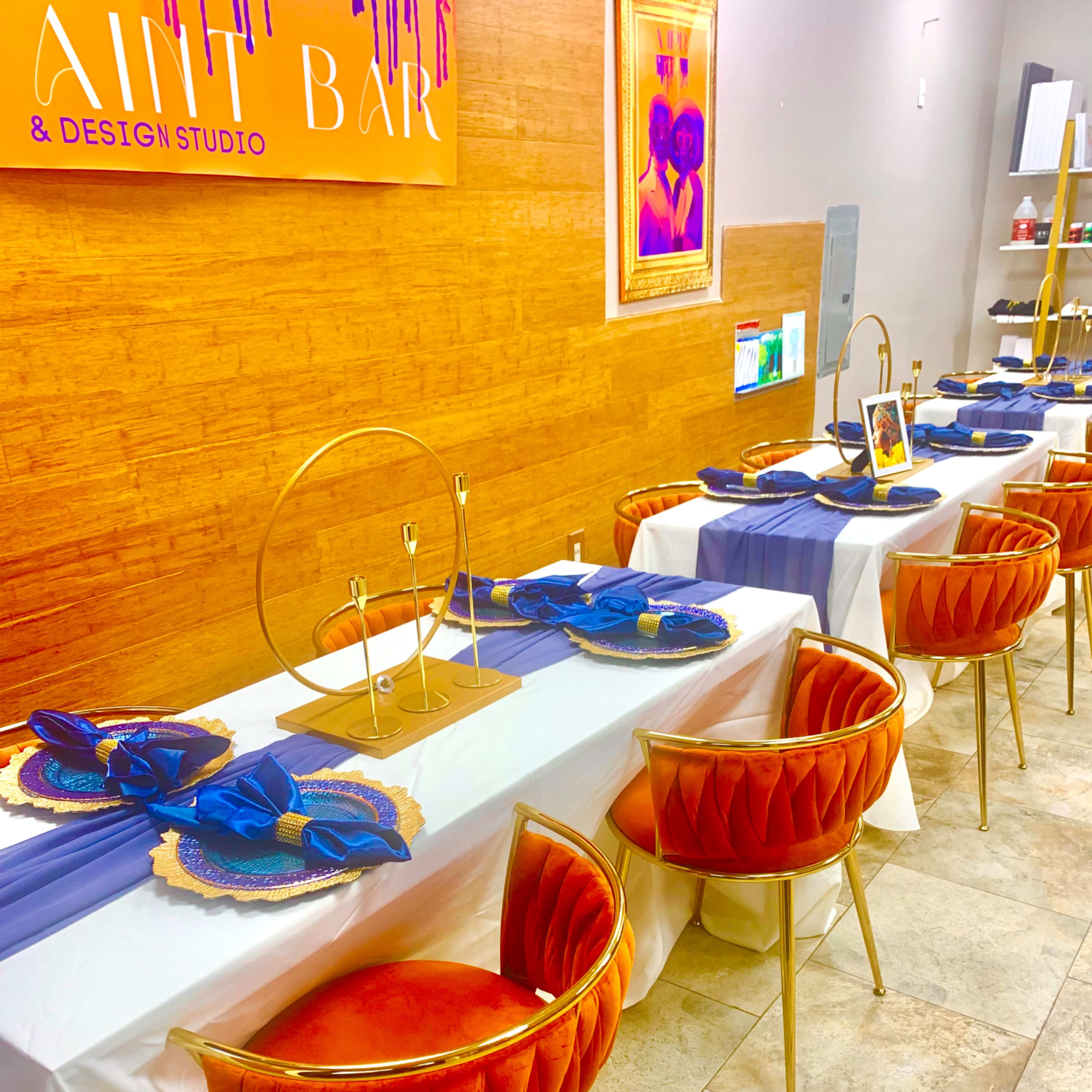 A dining setup with a long table covered in white and purple tablecloths, adorned with gold and blue tableware and velvet chairs.