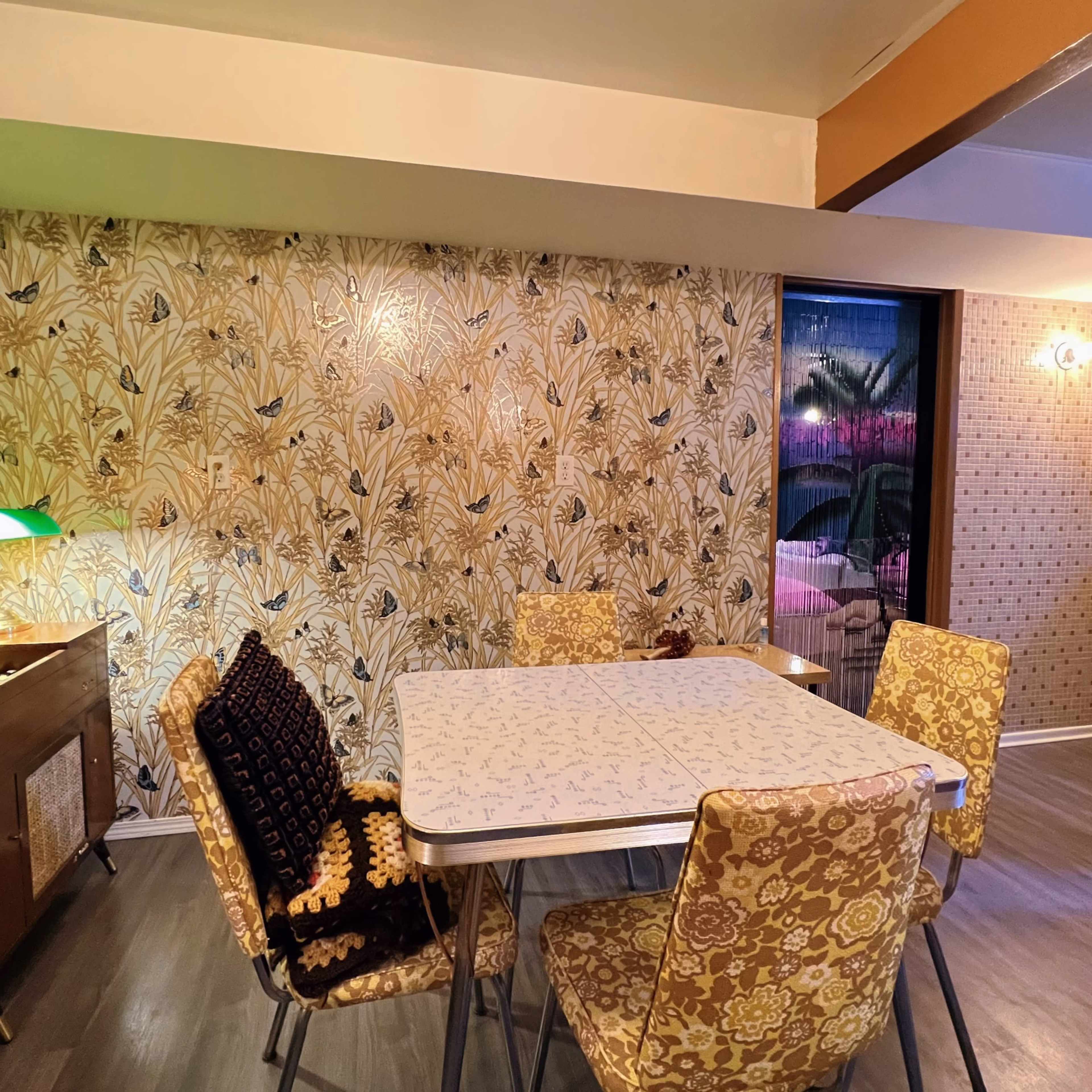 A dining area with a patterned wallpaper, a table surrounded by six chairs, and a sideboard with a green lamp.