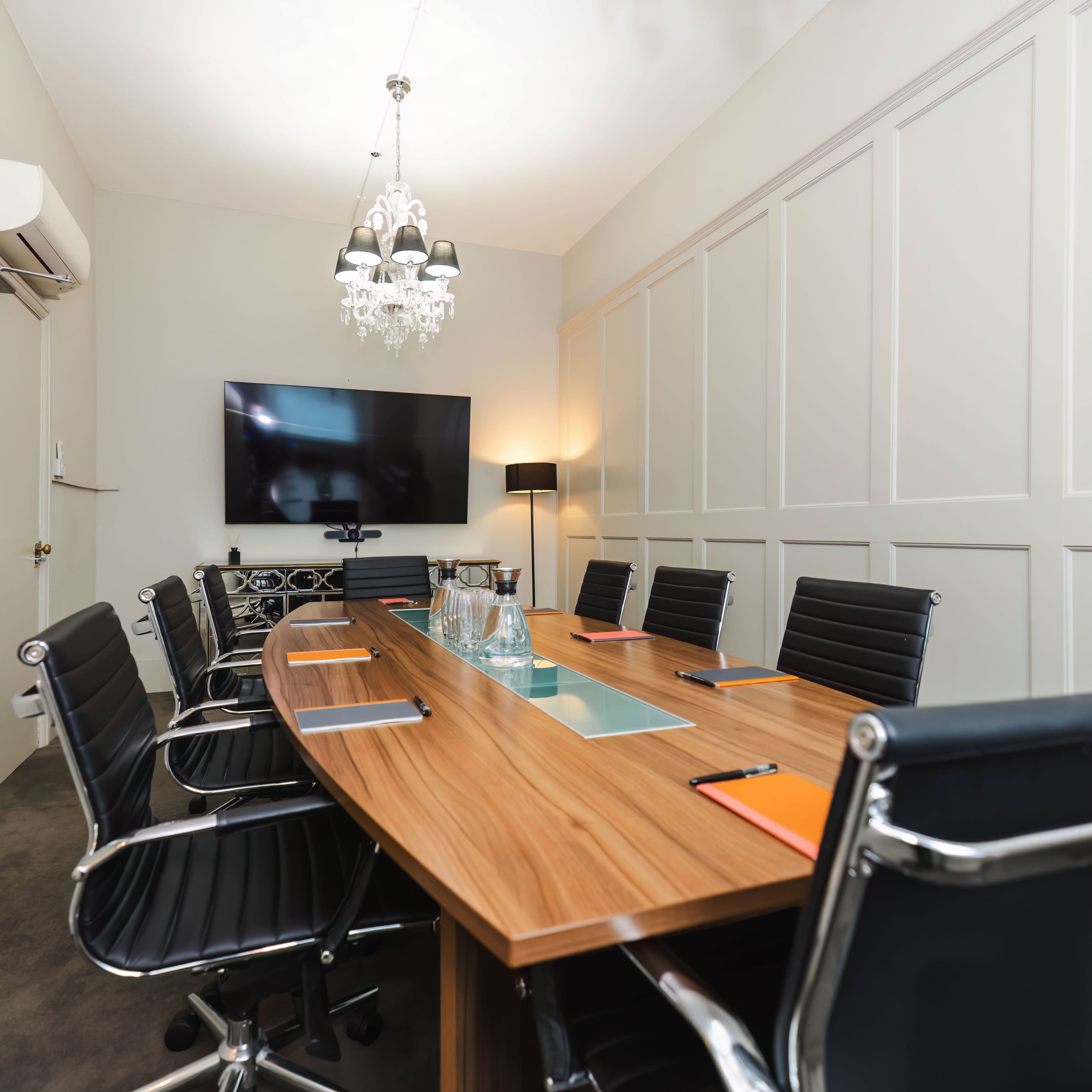 A modern conference room with a long wooden table, black leather chairs, a large TV mounted on the wall, and minimalist decor.