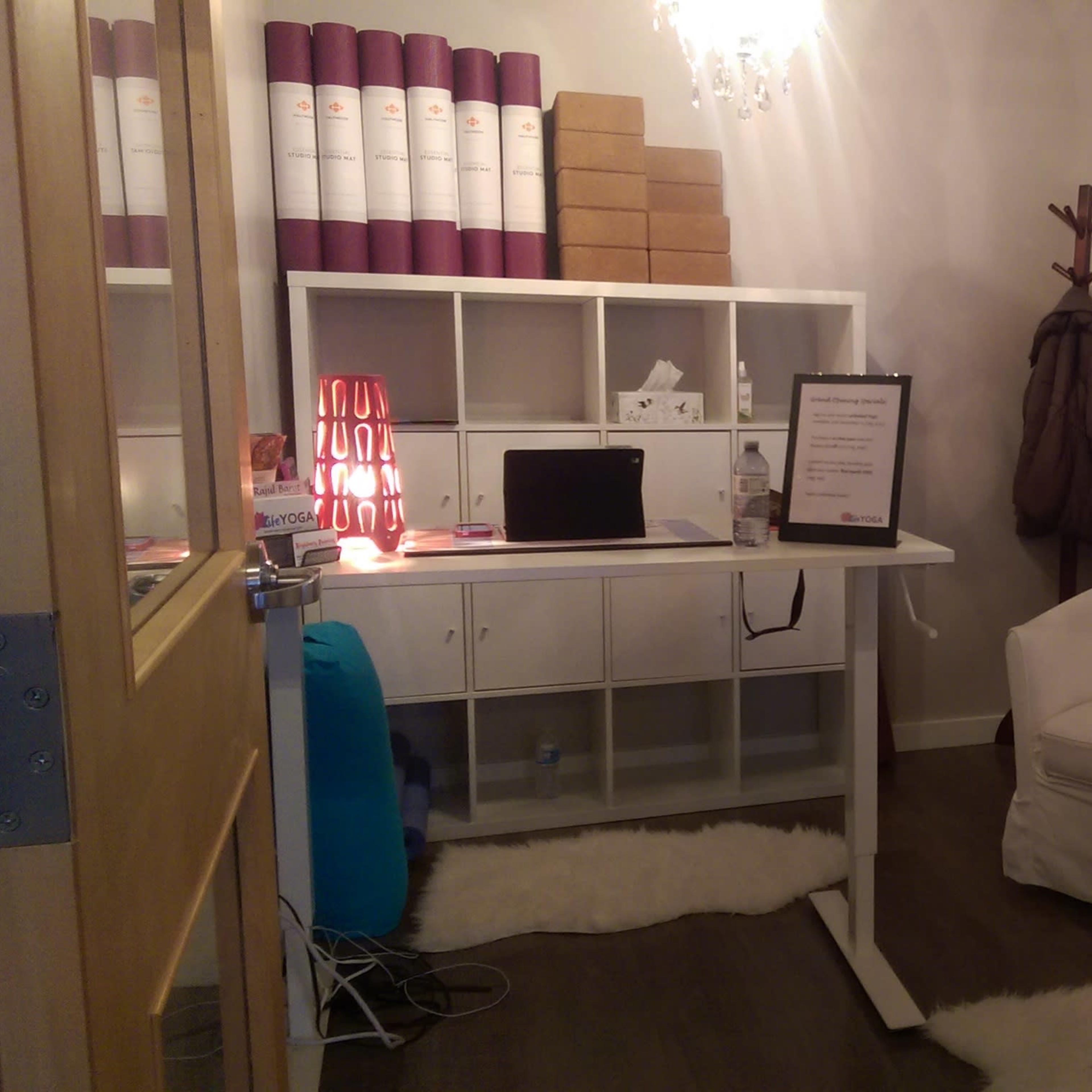 A small office space with a white desk, a chair, and storage boxes on shelves, illuminated by a chandelier.