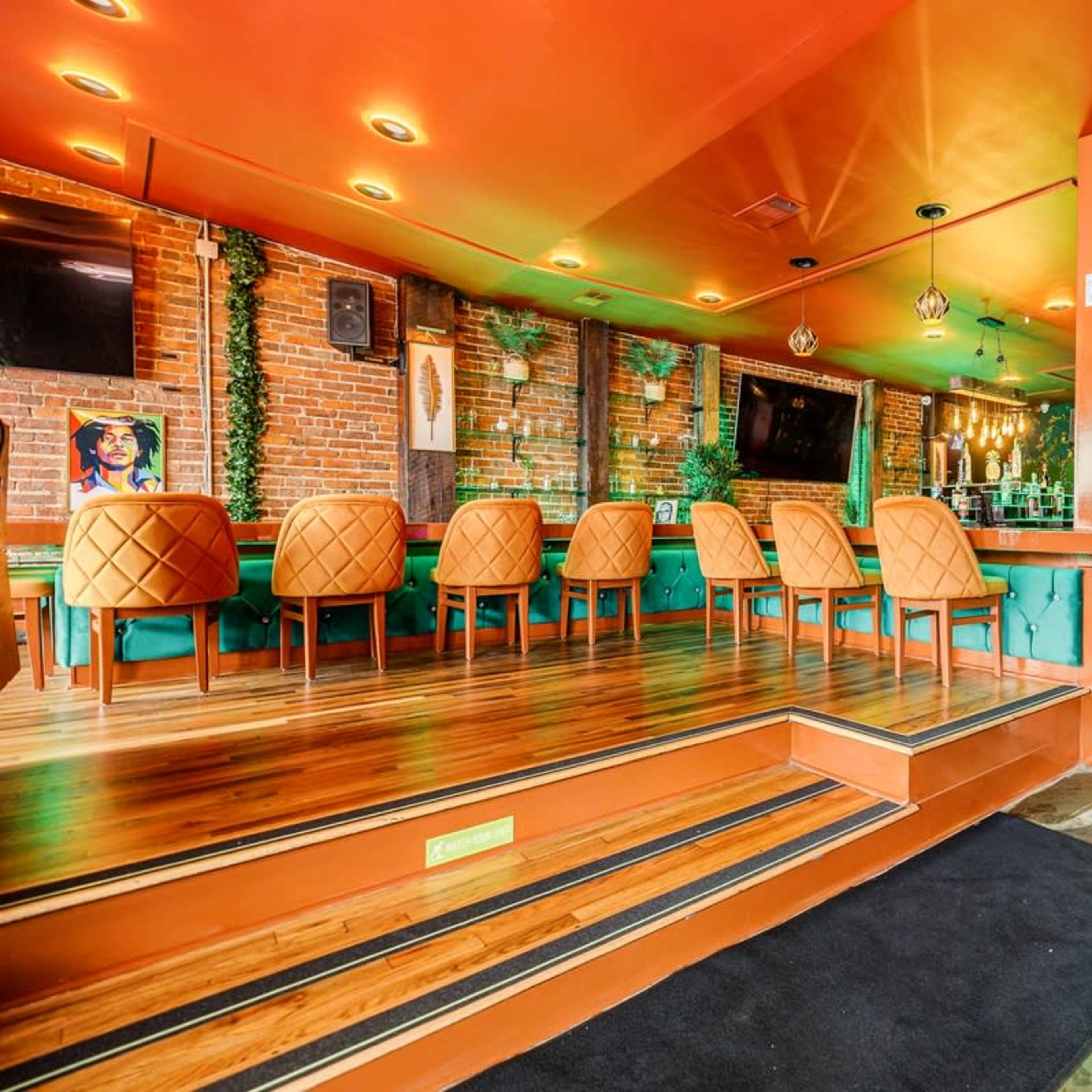 A vibrant bar interior with orange walls, green seating, and wooden flooring, featuring a mix of high and low seating arrangements.