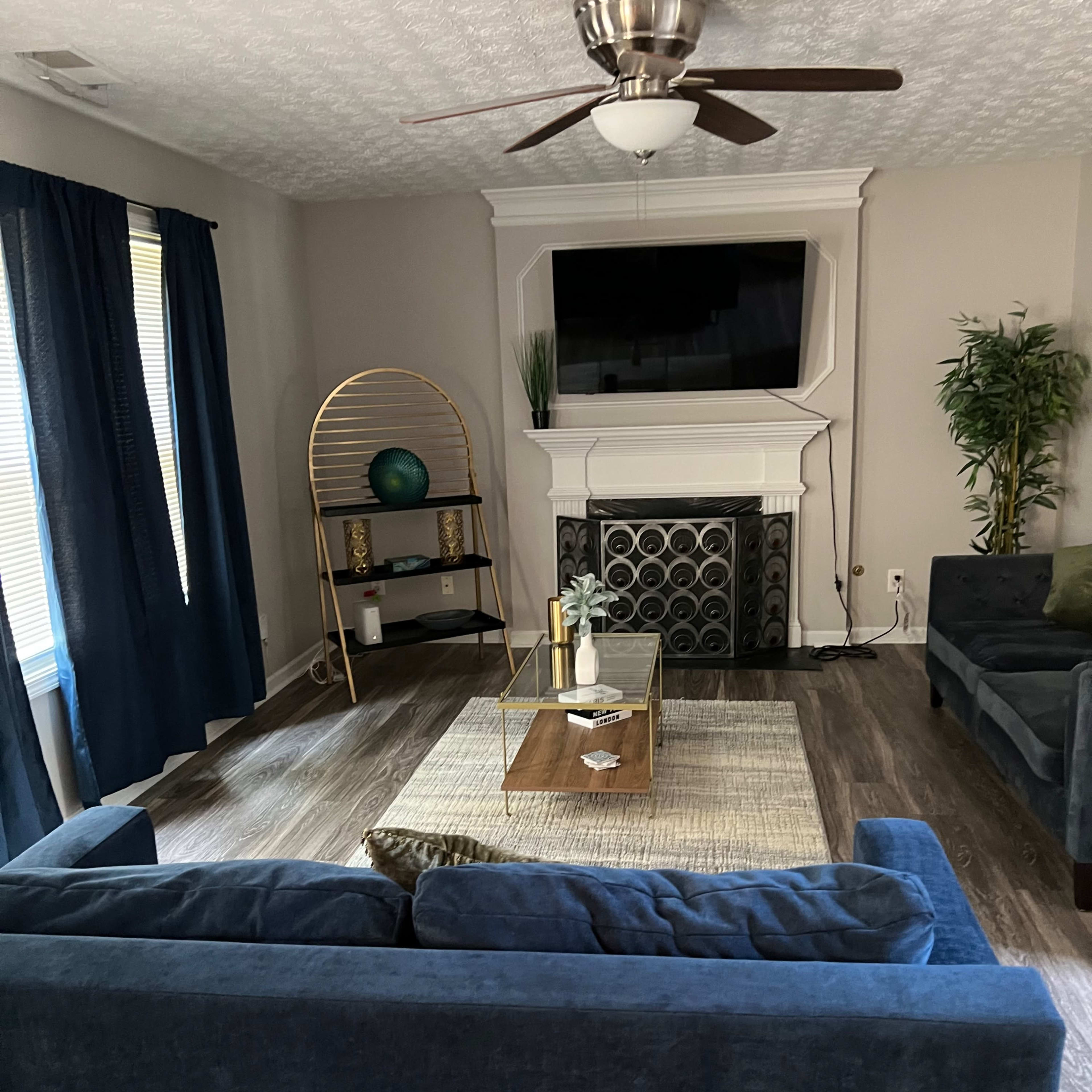 The living room features a blue sofa, a coffee table on a textured rug, and a media console beneath a mounted TV, with large windows dressed in navy curtains.