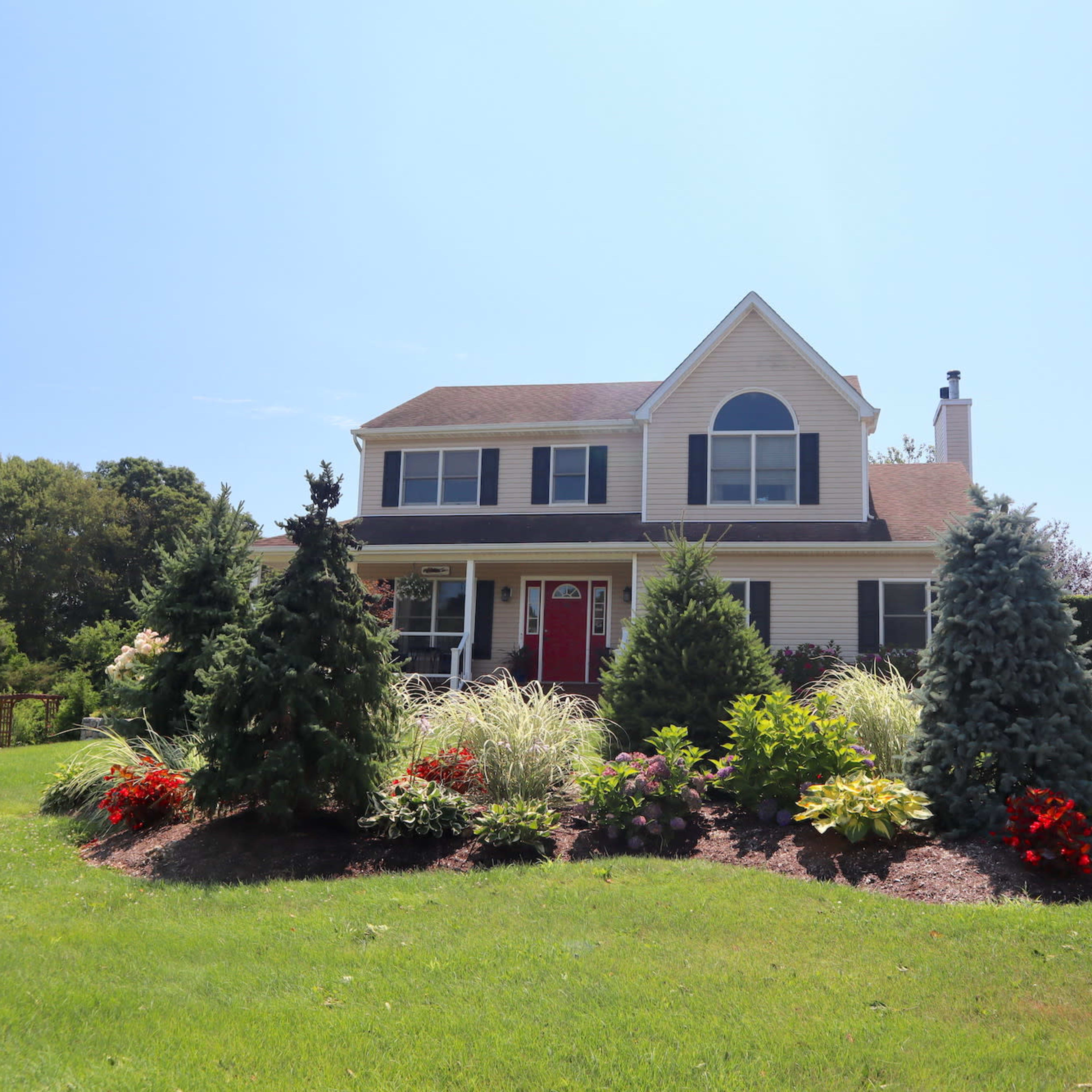 A two-story house with a red door is surrounded by well-maintained landscaping featuring flowering plants and evergreen trees.