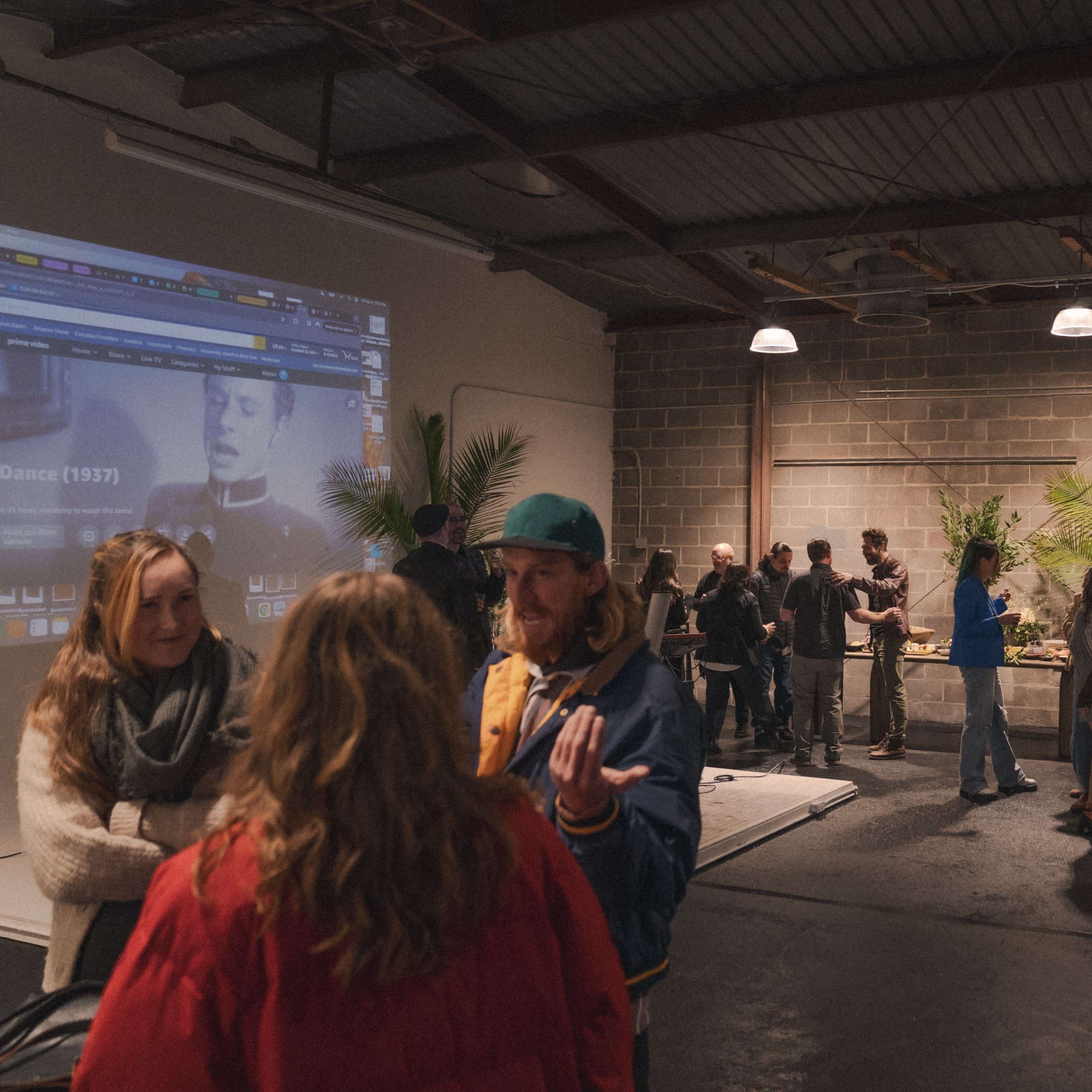 A group of people gathers in an industrial-style space with a large screen displaying video content, while others engage in conversation amidst plants and minimal furnishings.