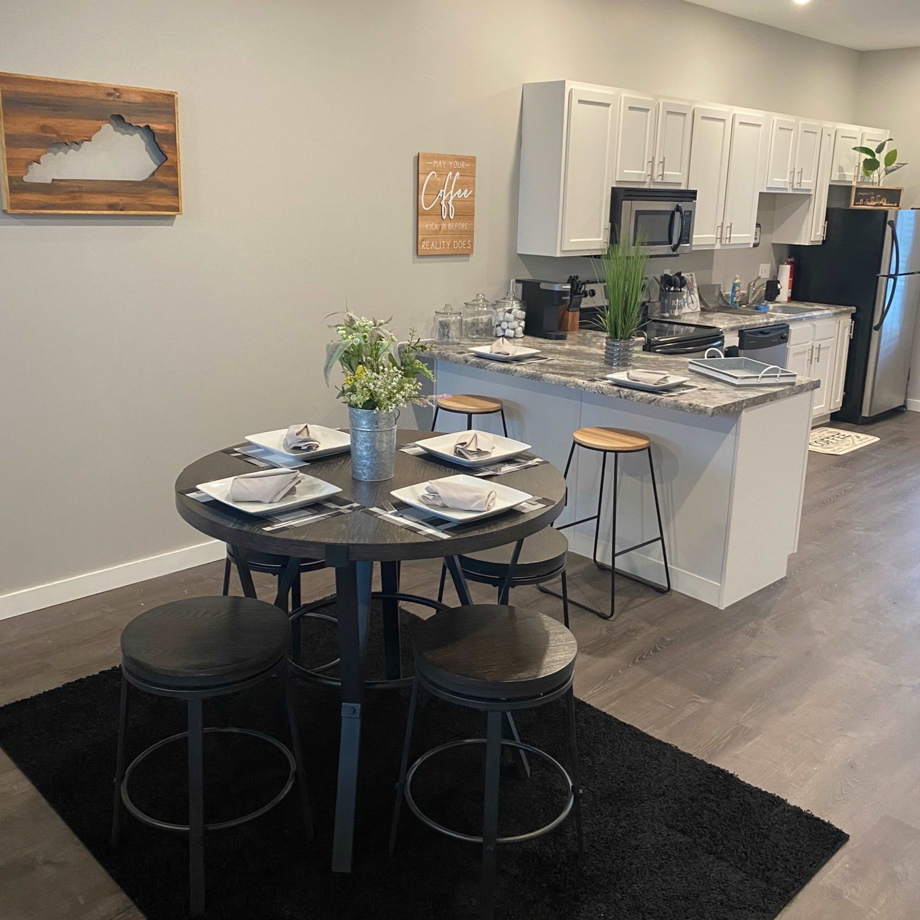 A modern dining area with a round table set for four, surrounded by black stools, adjacent to a kitchen with white cabinets and appliances.
