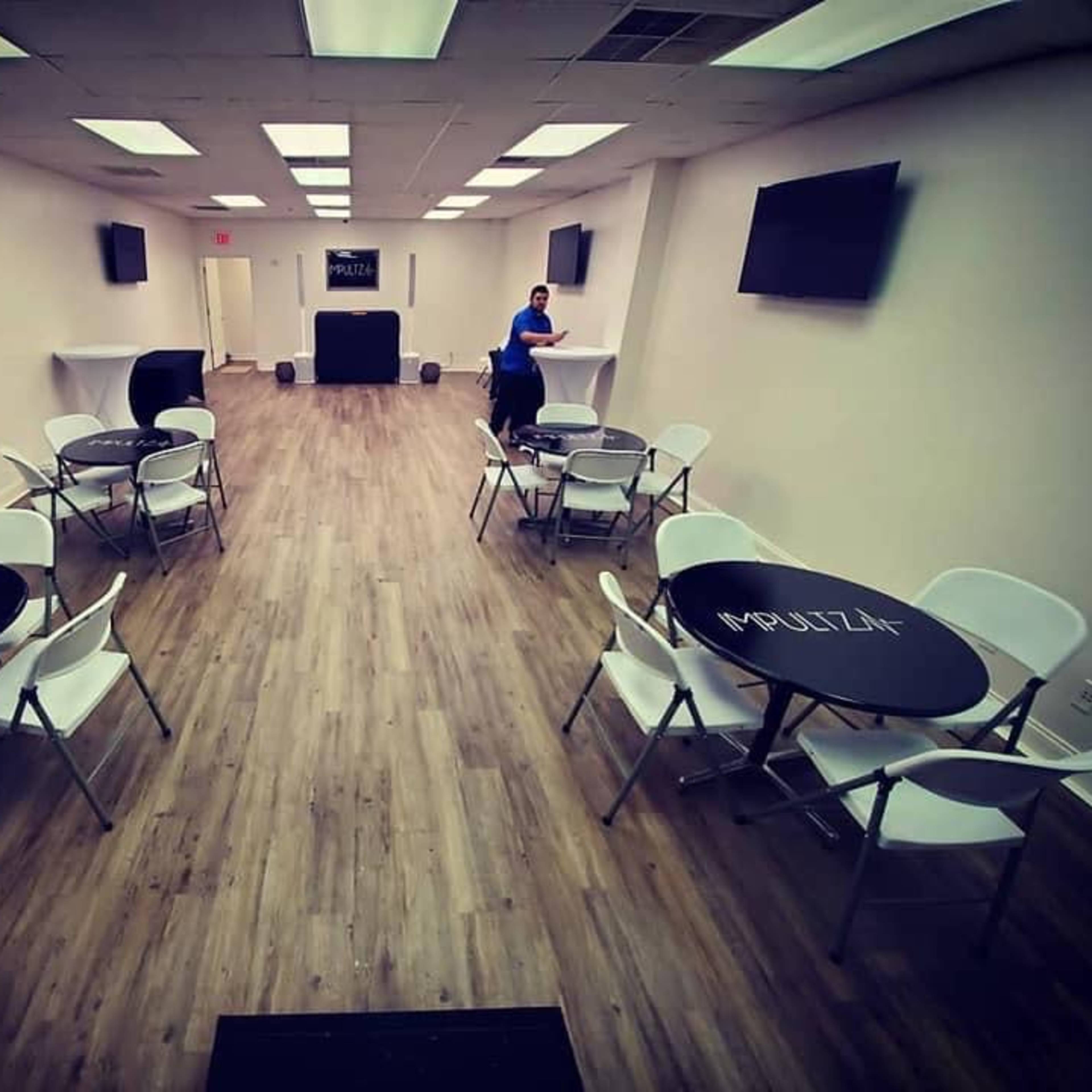 An empty event space with several round tables and chairs arranged on a wooden floor, illuminated by overhead lights.