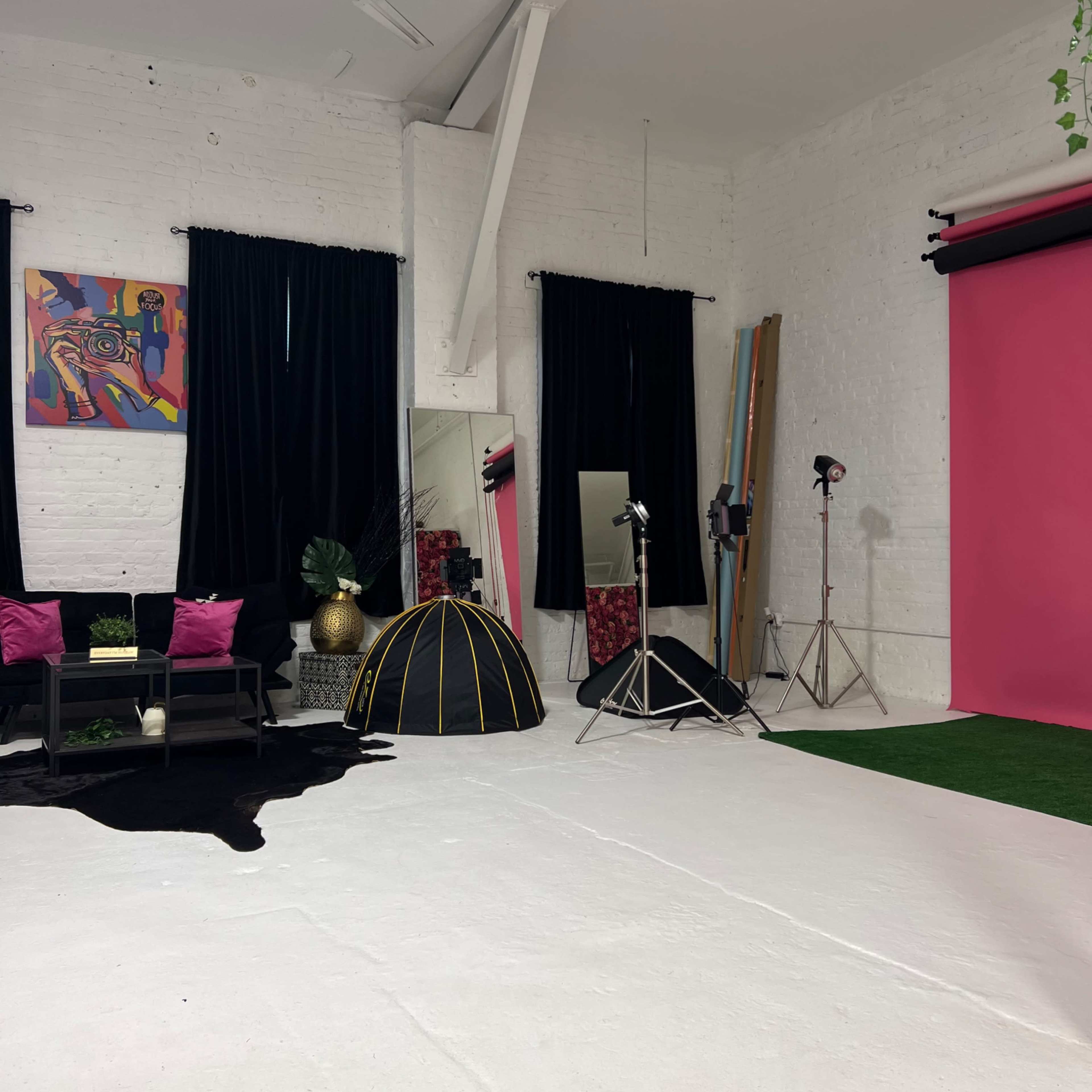 A photography studio with a pink backdrop, black curtains, a seating area, and various lighting equipment.