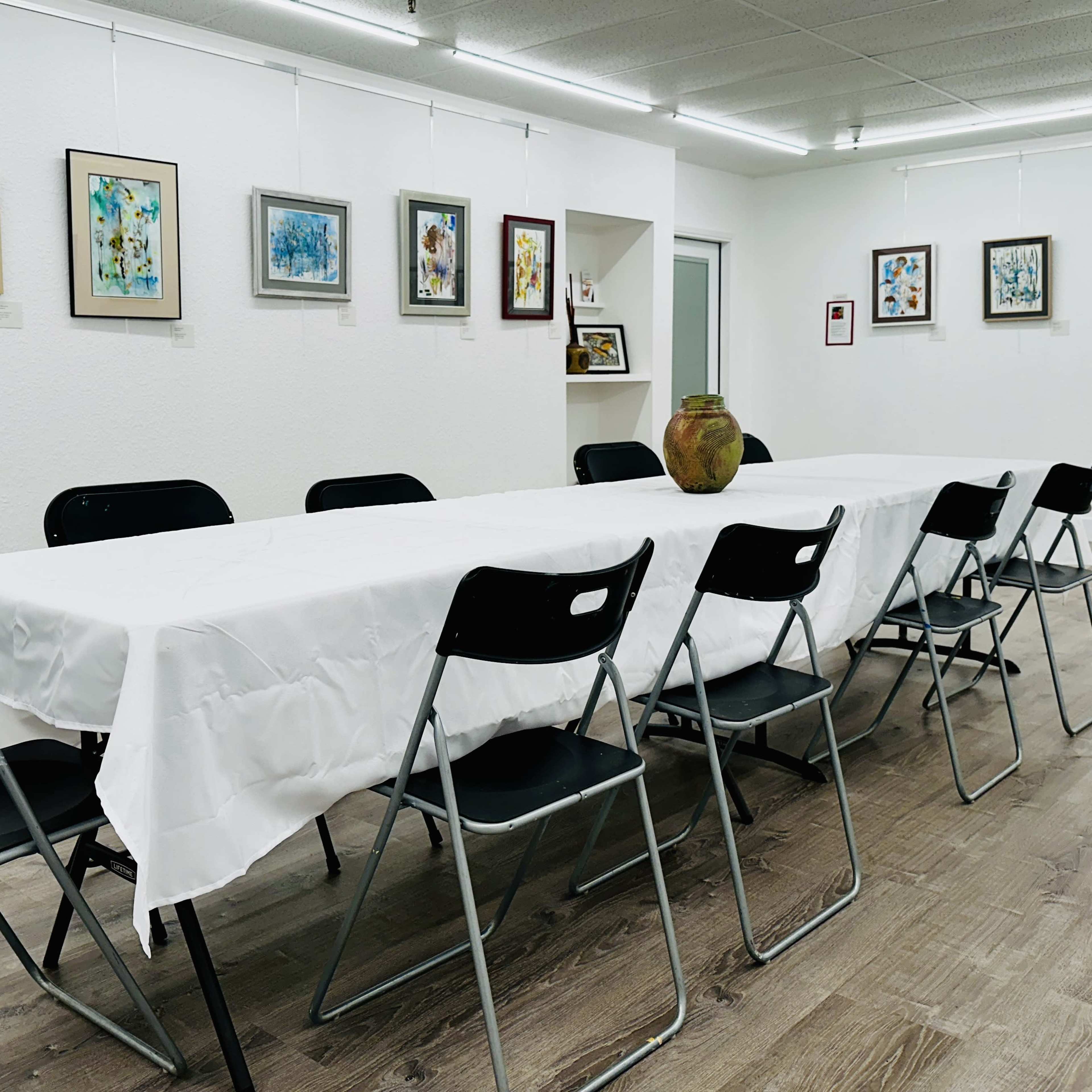 A spacious art gallery with white walls, featuring a long table covered with a white tablecloth and surrounded by black folding chairs, while various framed artworks are hung on the walls.