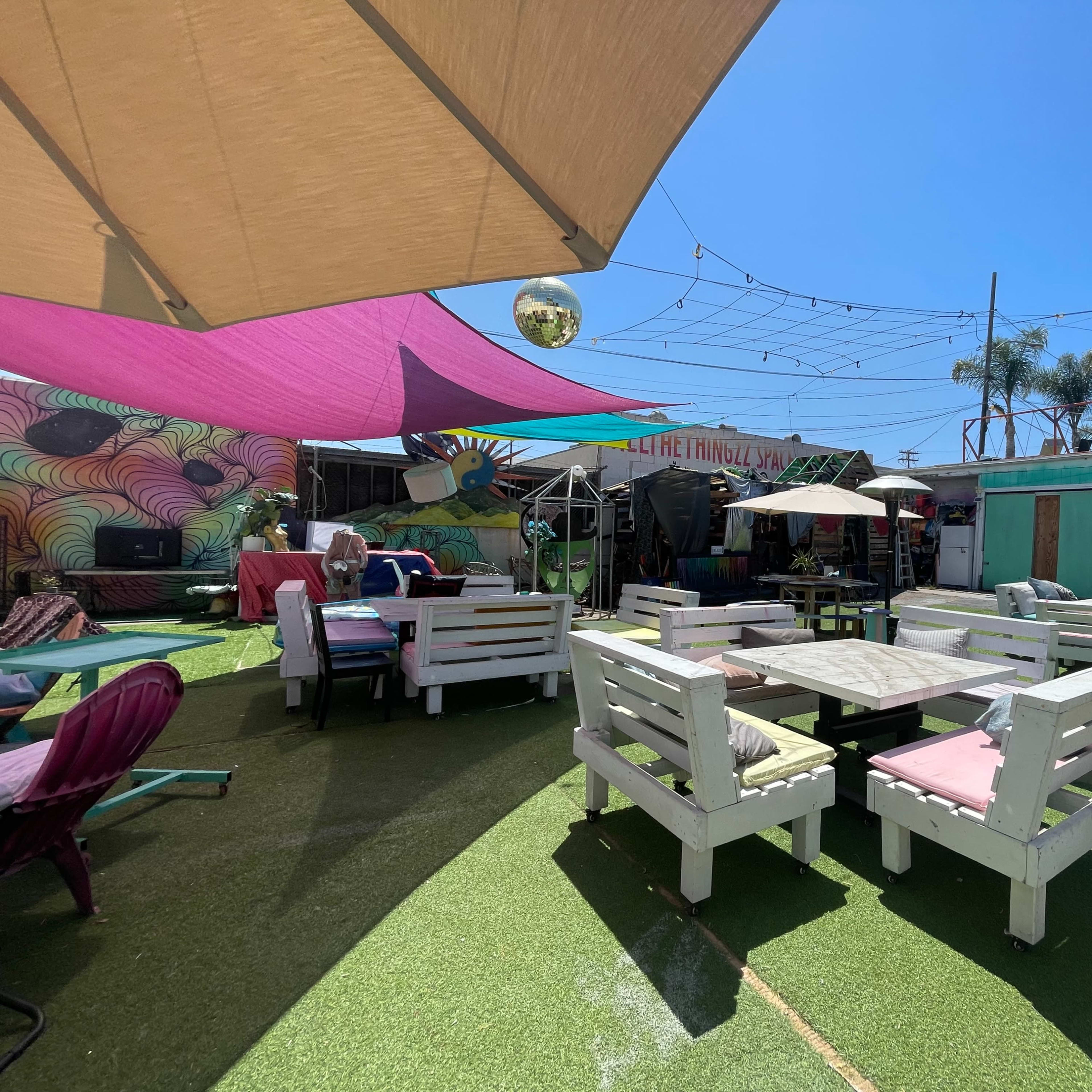 A vibrant outdoor seating area with colorful umbrellas and wooden tables surrounded by decorative murals and greenery.