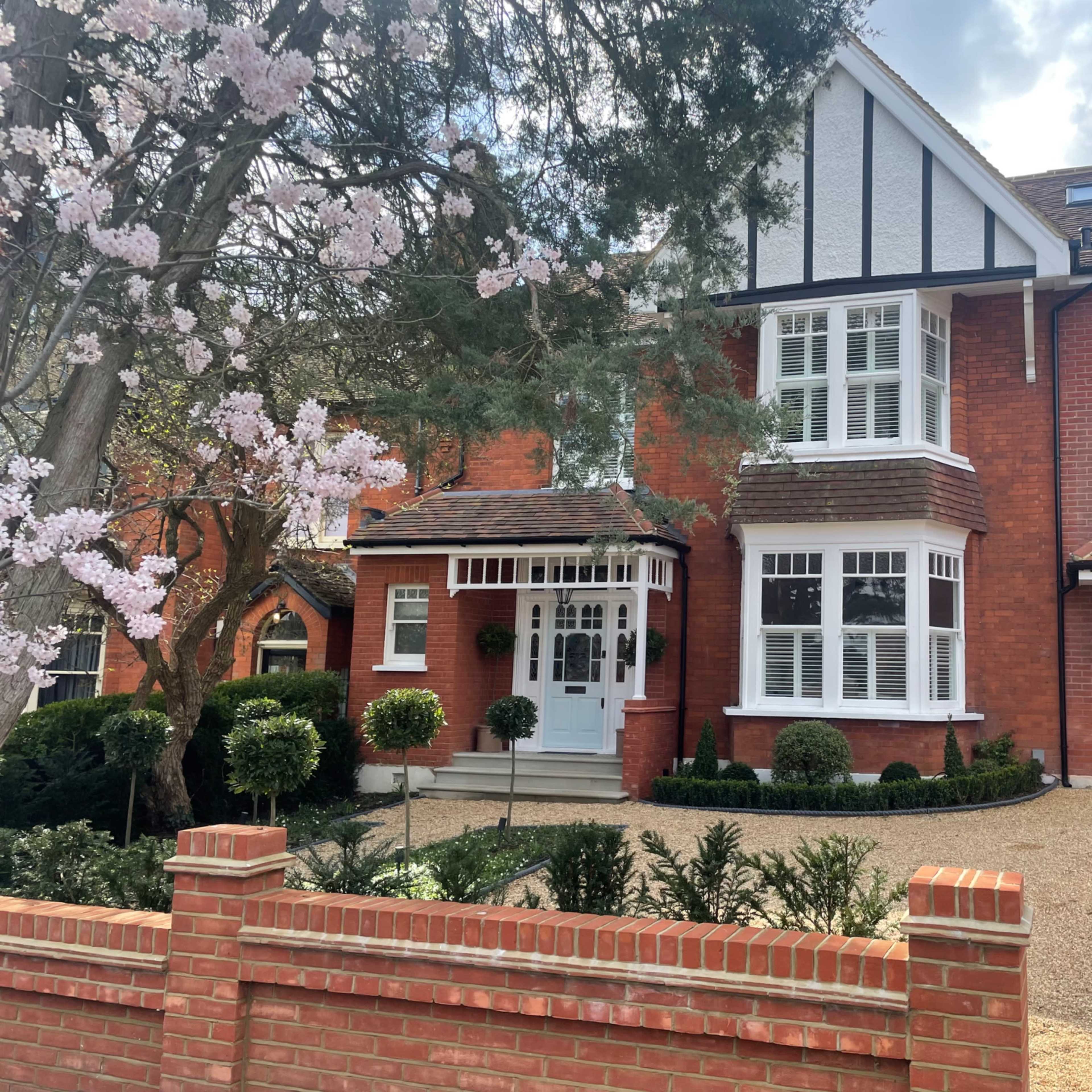 A red-brick house with a gabled roof, large windows, and a neatly landscaped front yard featuring a flowering tree and a gravel driveway.