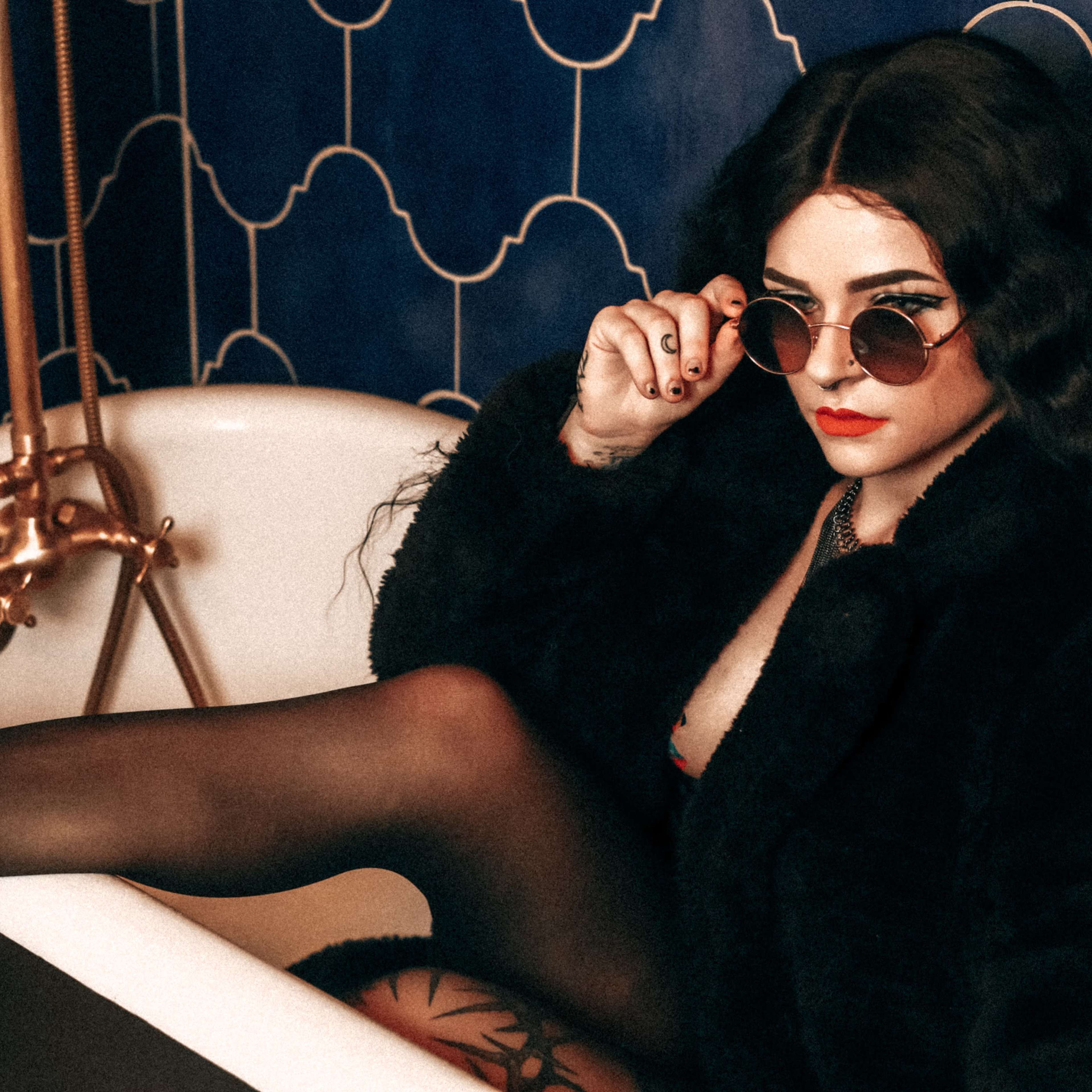 A woman in sunglasses lounges in a vintage bathtub, wearing a black fur coat and thigh-high stockings against a blue tiled backdrop.