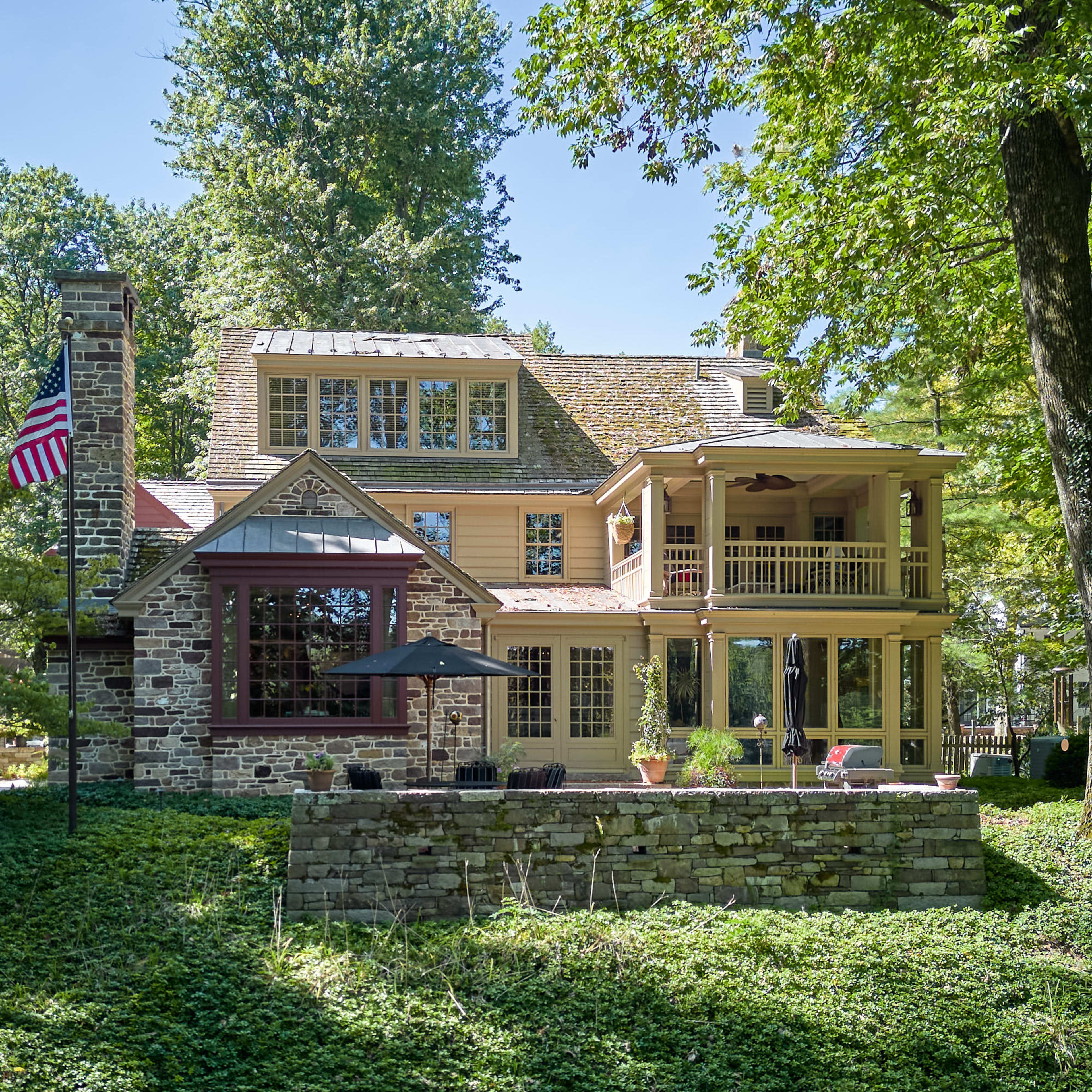 A large, multi-story stone and wood house with a porch, surrounded by trees and greenery.