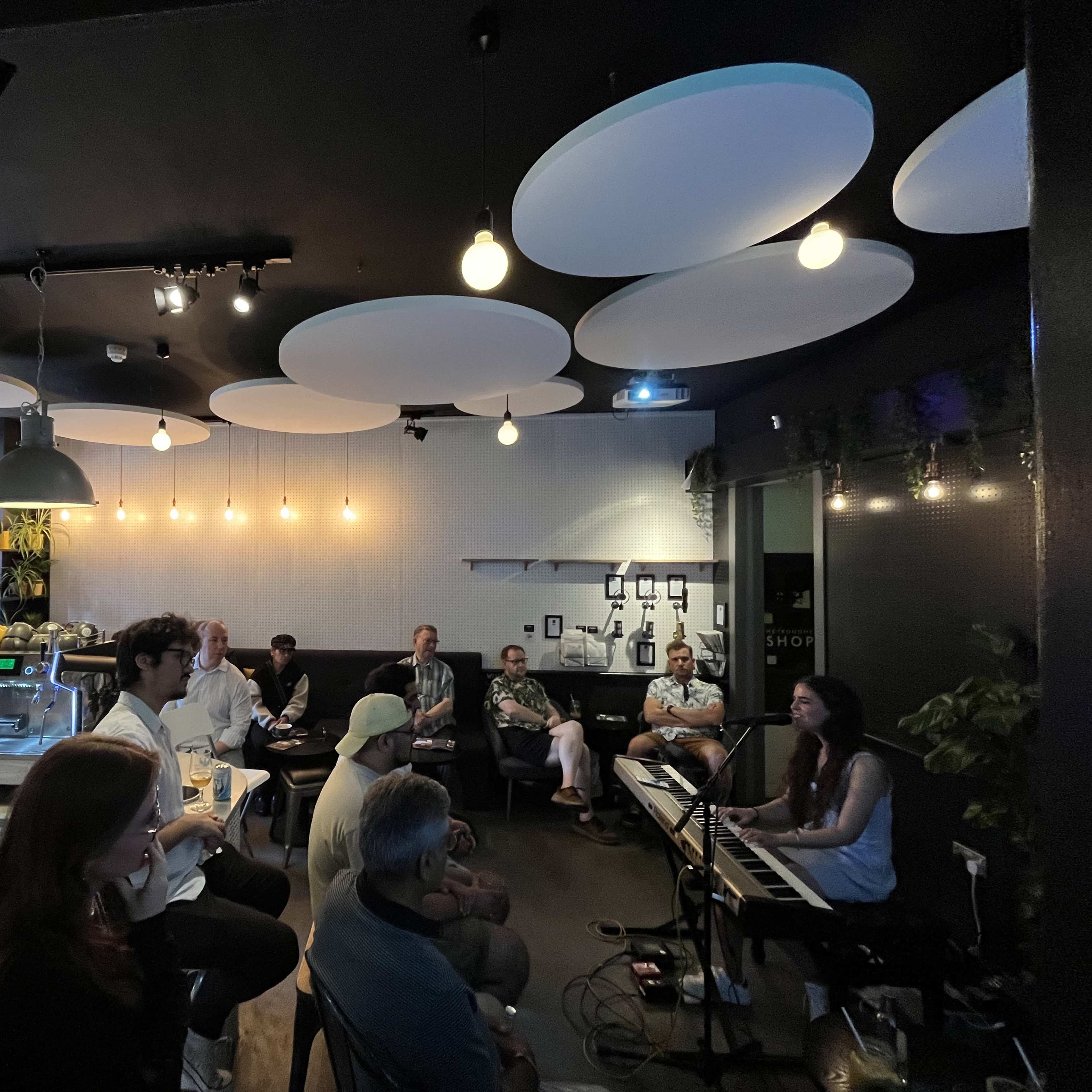 A small crowd listens to a musician playing a piano in a cozy café adorned with modern lighting and décor.
