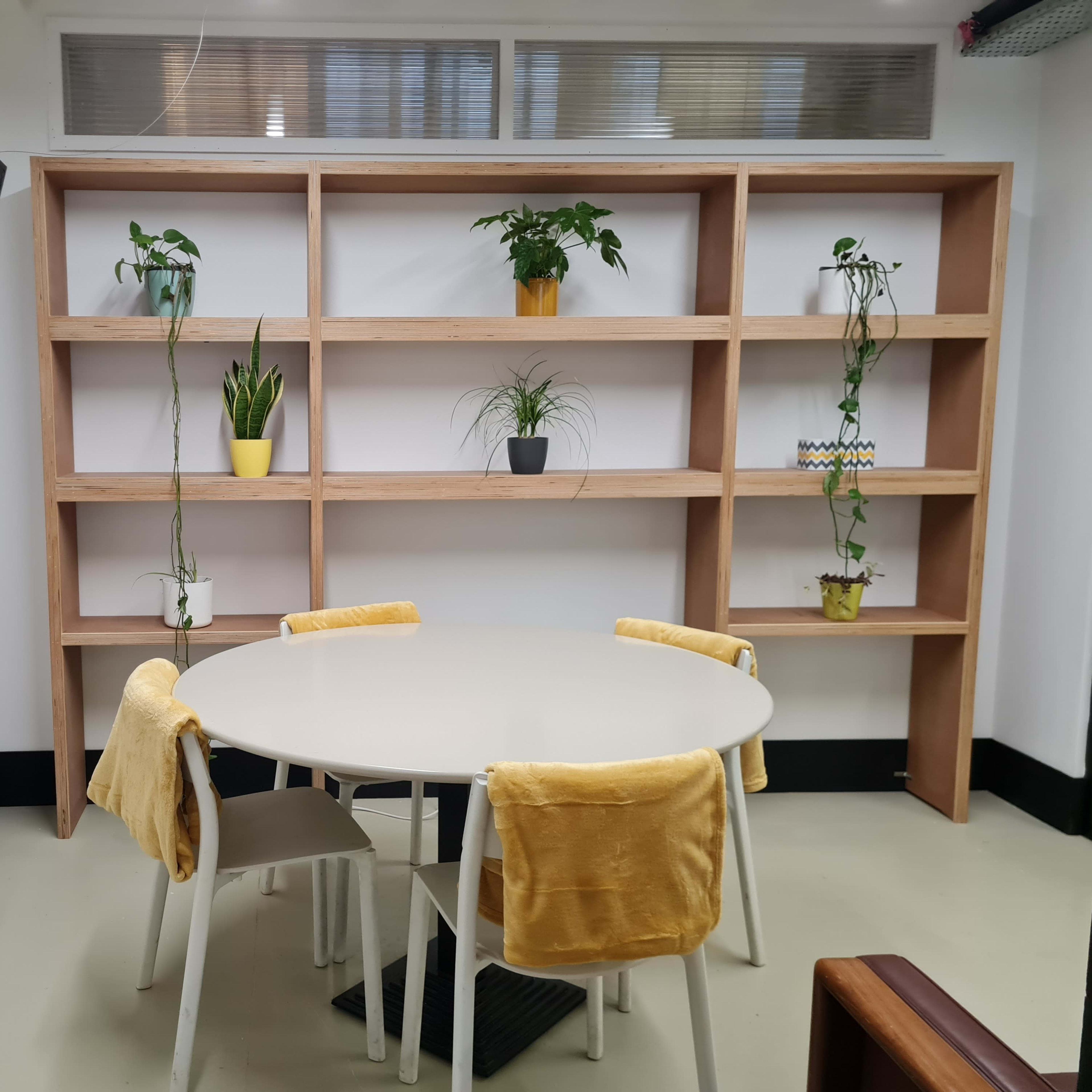 A circular table surrounded by four chairs is positioned in front of a wooden shelf with potted plants and a wall-mounted television.
