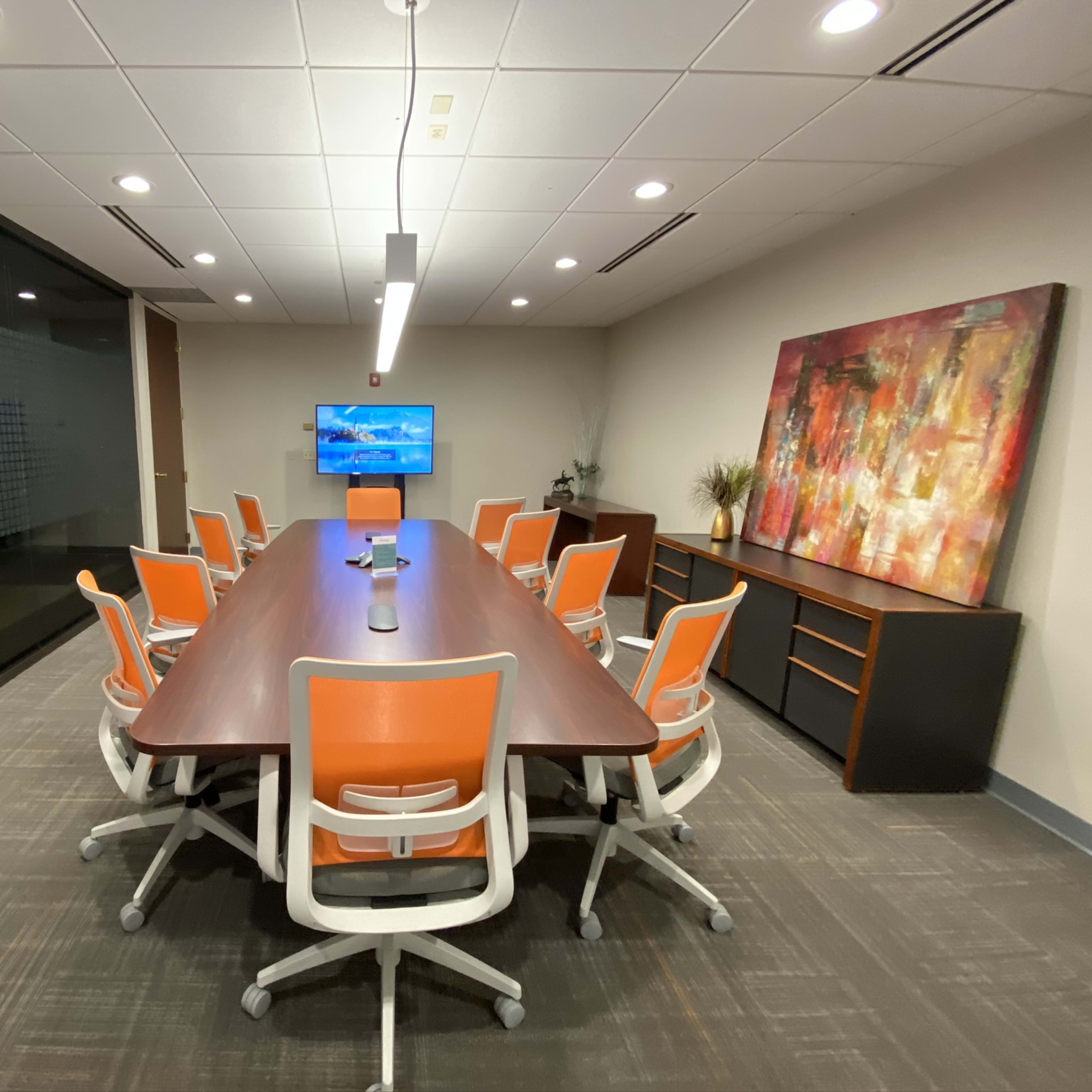 A modern conference room features a large rectangular table surrounded by orange and white chairs, with a TV mounted on the wall and a colorful abstract painting.