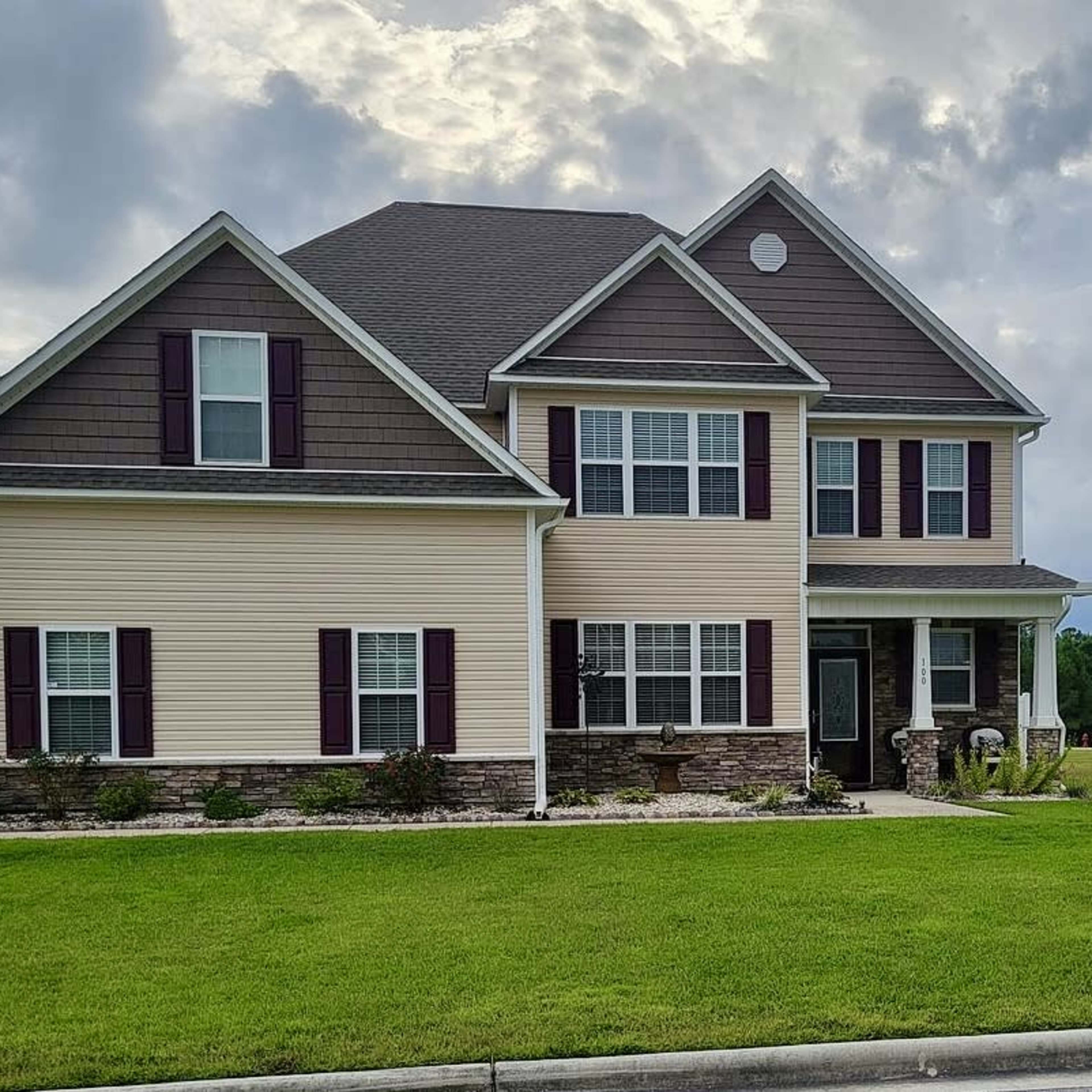 A two-story house with a combination of beige siding and dark brown accents sits on a green lawn under a cloudy sky.