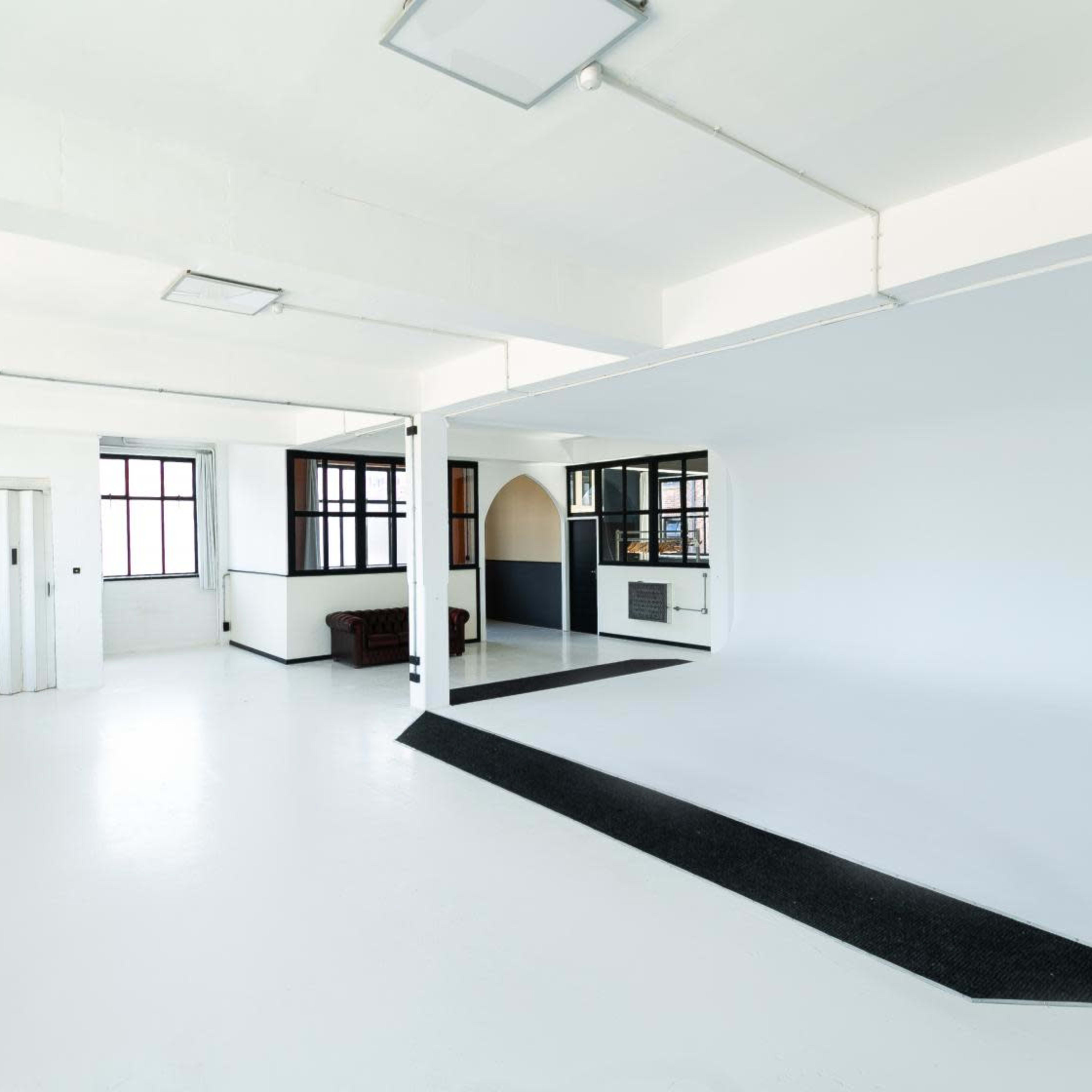 A spacious, empty interior with white walls, a polished floor, and large windows, featuring a small partitioned area in the center.