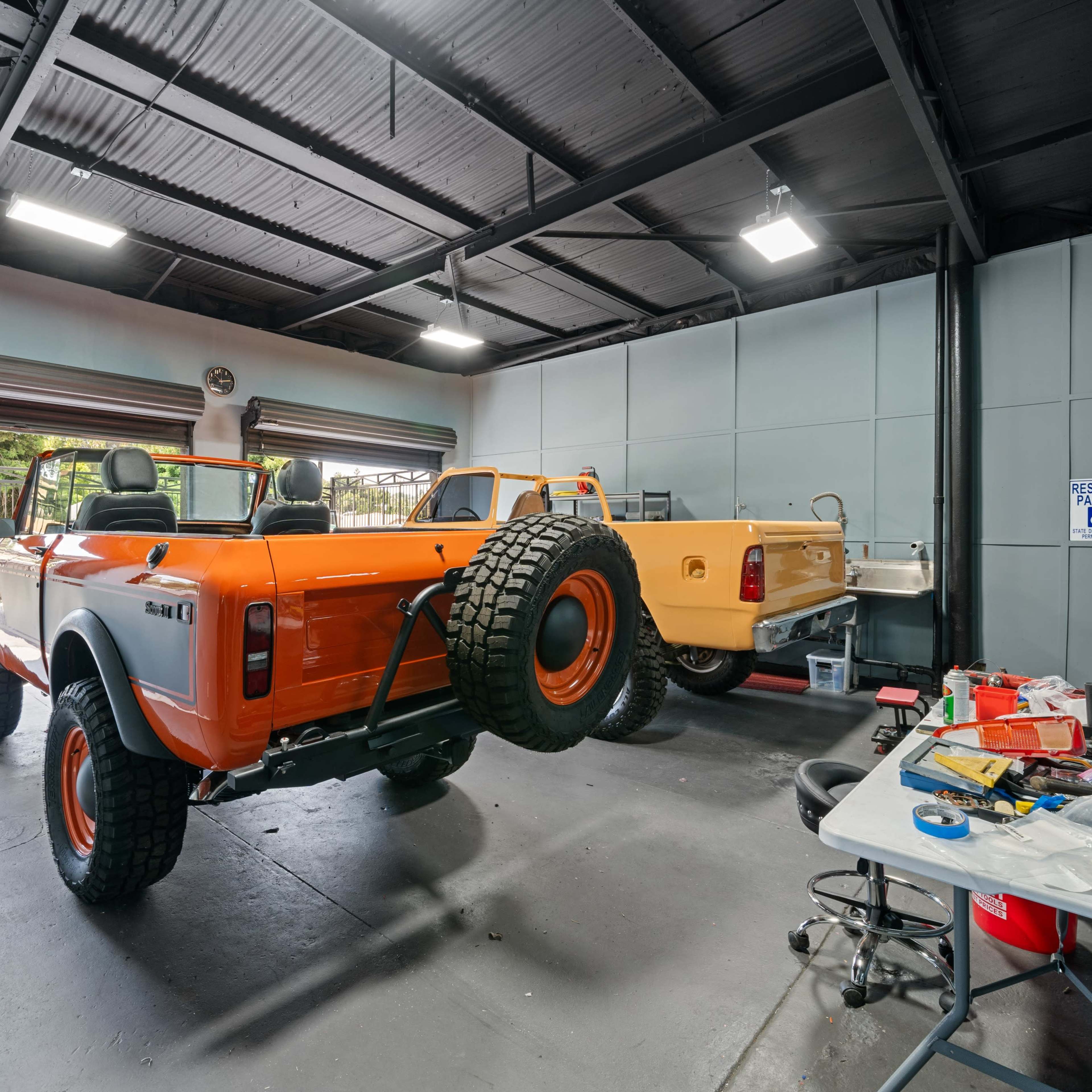 An orange off-road vehicle in a garage alongside a yellow vehicle, with various tools and equipment scattered on a table.