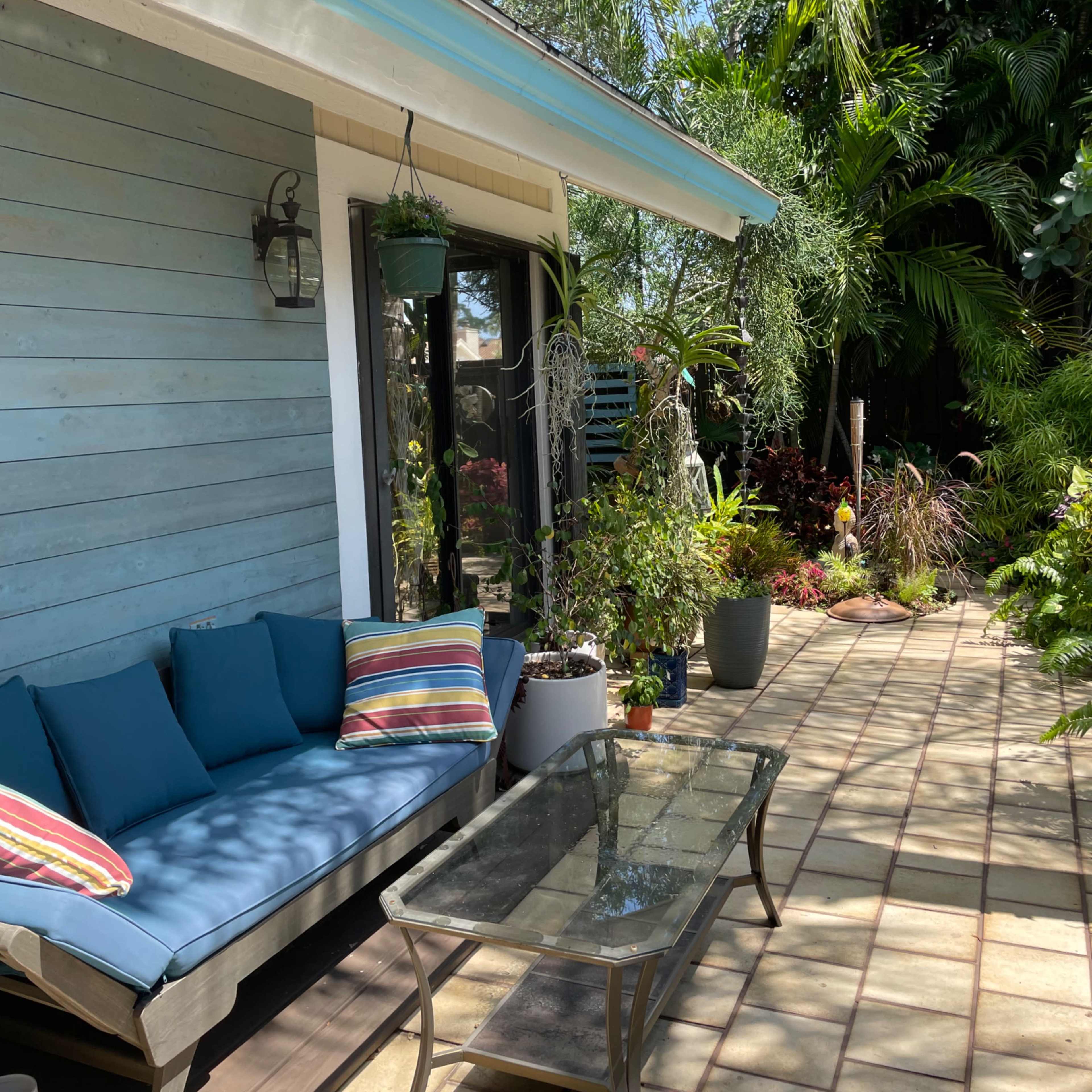 A patio with a couch, striped cushions, and a glass coffee table is surrounded by lush greenery and potted plants.