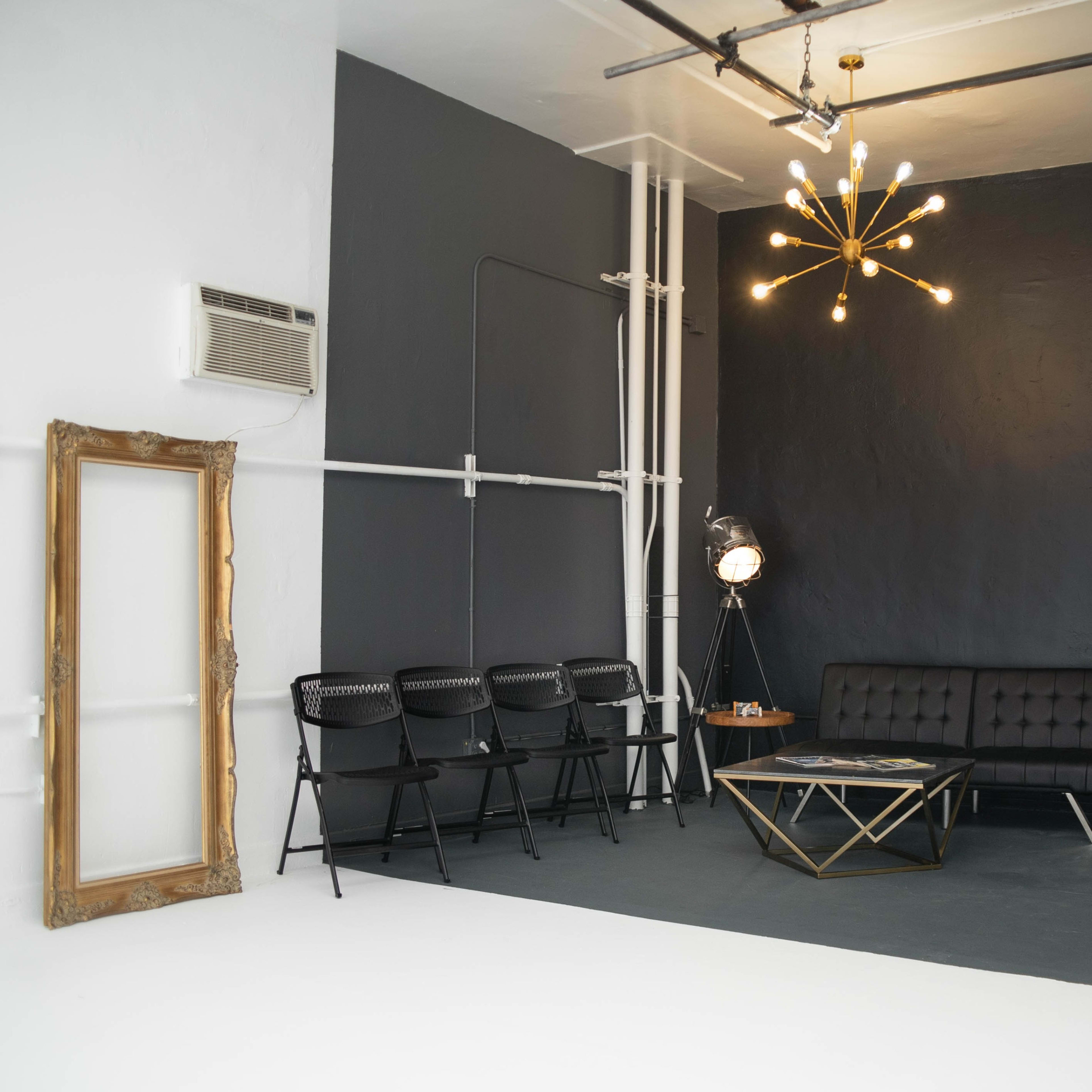 A minimalist interior space featuring a black wall, a tufted black sofa, a gold starburst chandelier, a small refrigerator, and a round glass coffee table surrounded by black chairs.