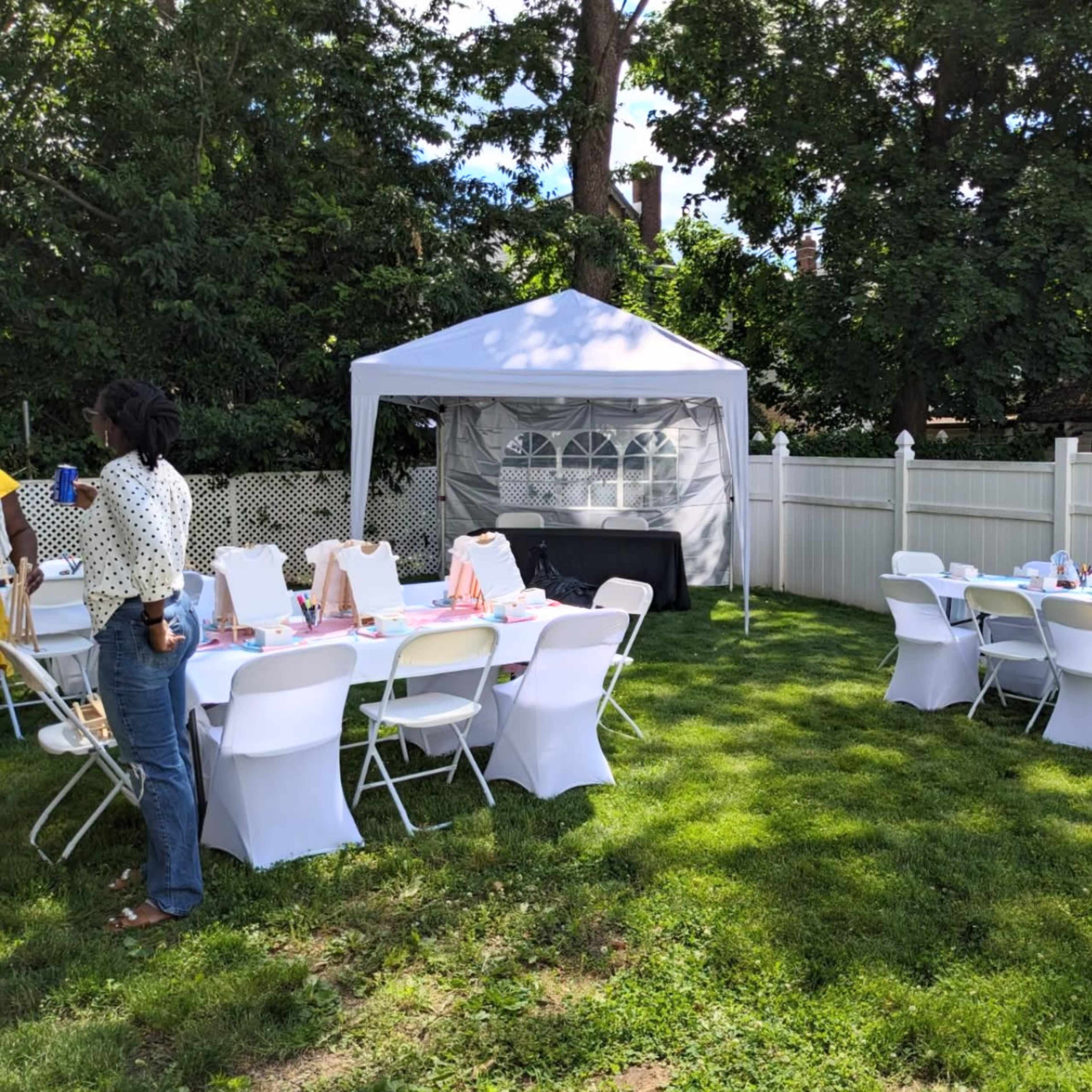 A backyard is set up for an outdoor gathering, featuring a white tent and several tables with chairs arranged on green grass.
