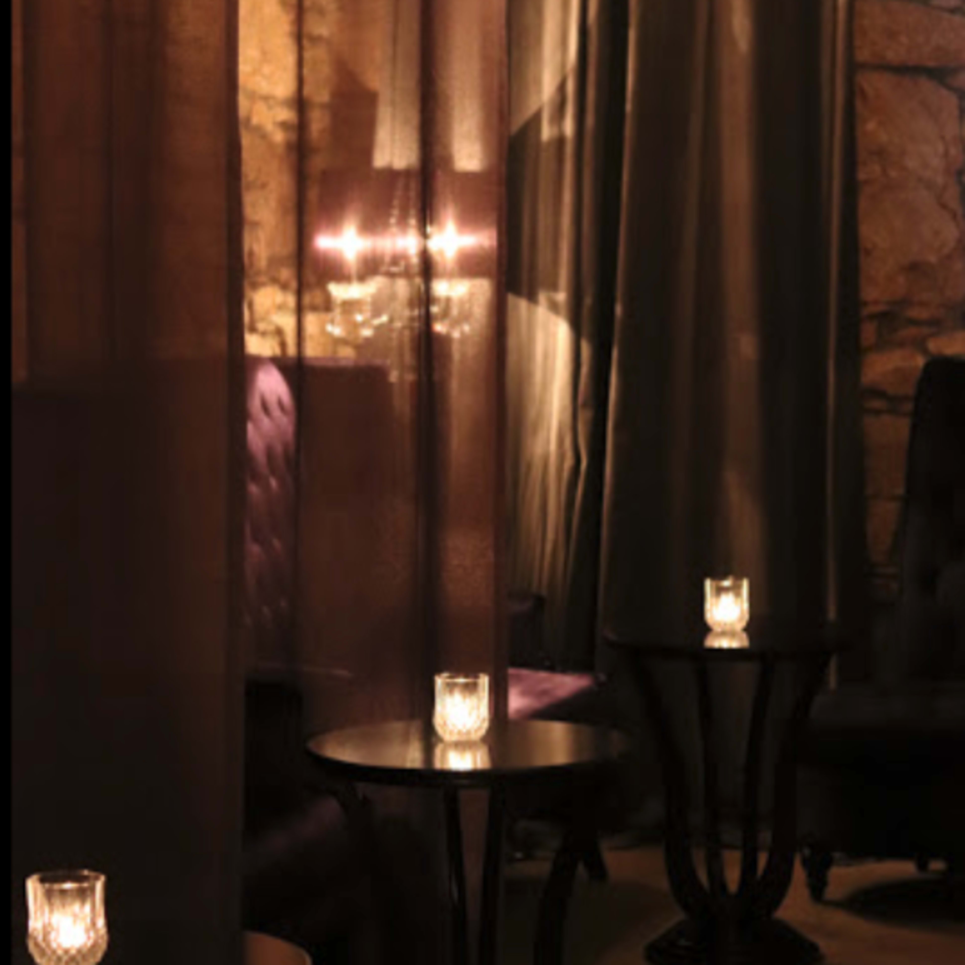 A dimly lit lounge with stone walls, sheer curtains, and candles on small tables.