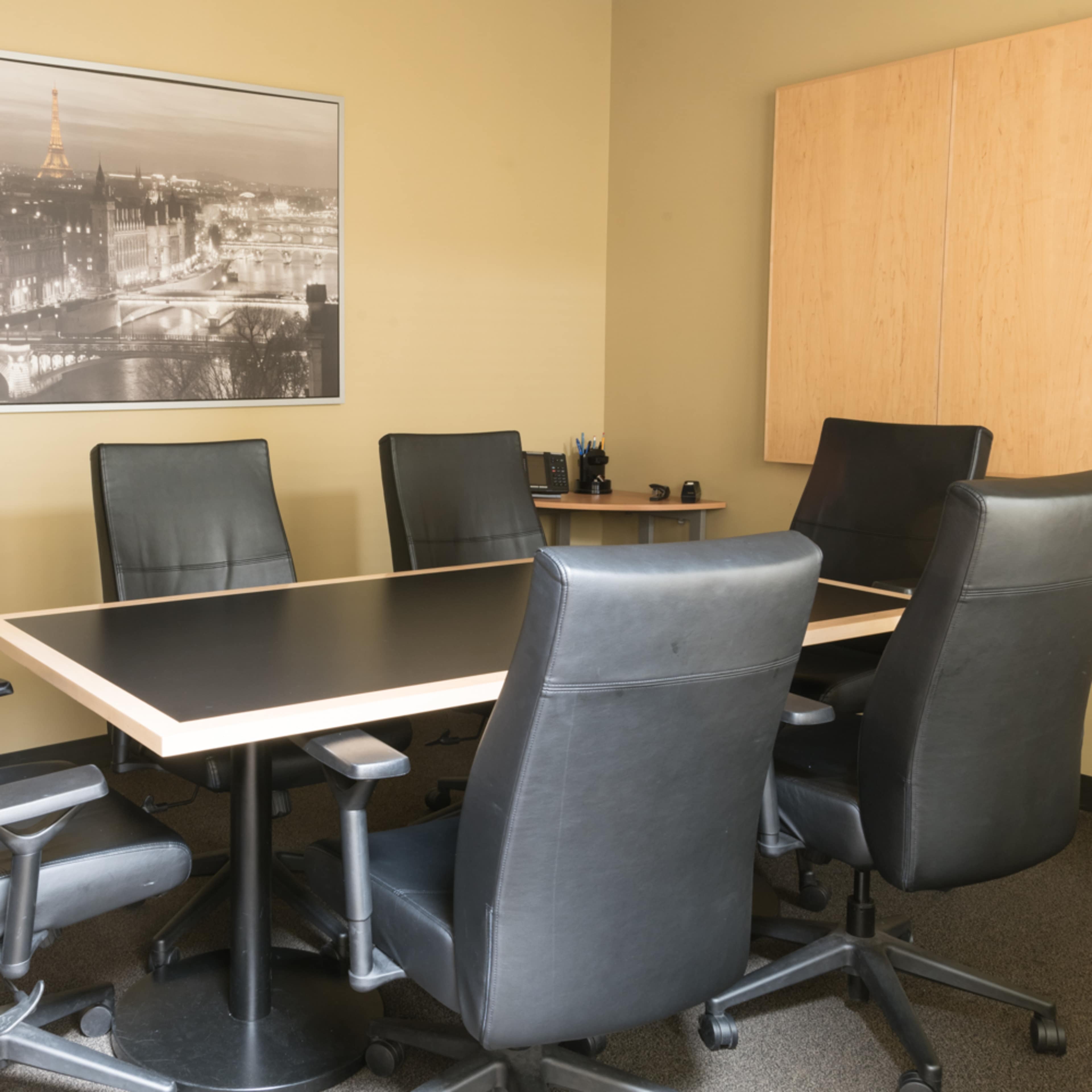 A conference room features a large rectangular table surrounded by six black leather chairs and a wall-mounted black-and-white photo of a cityscape.