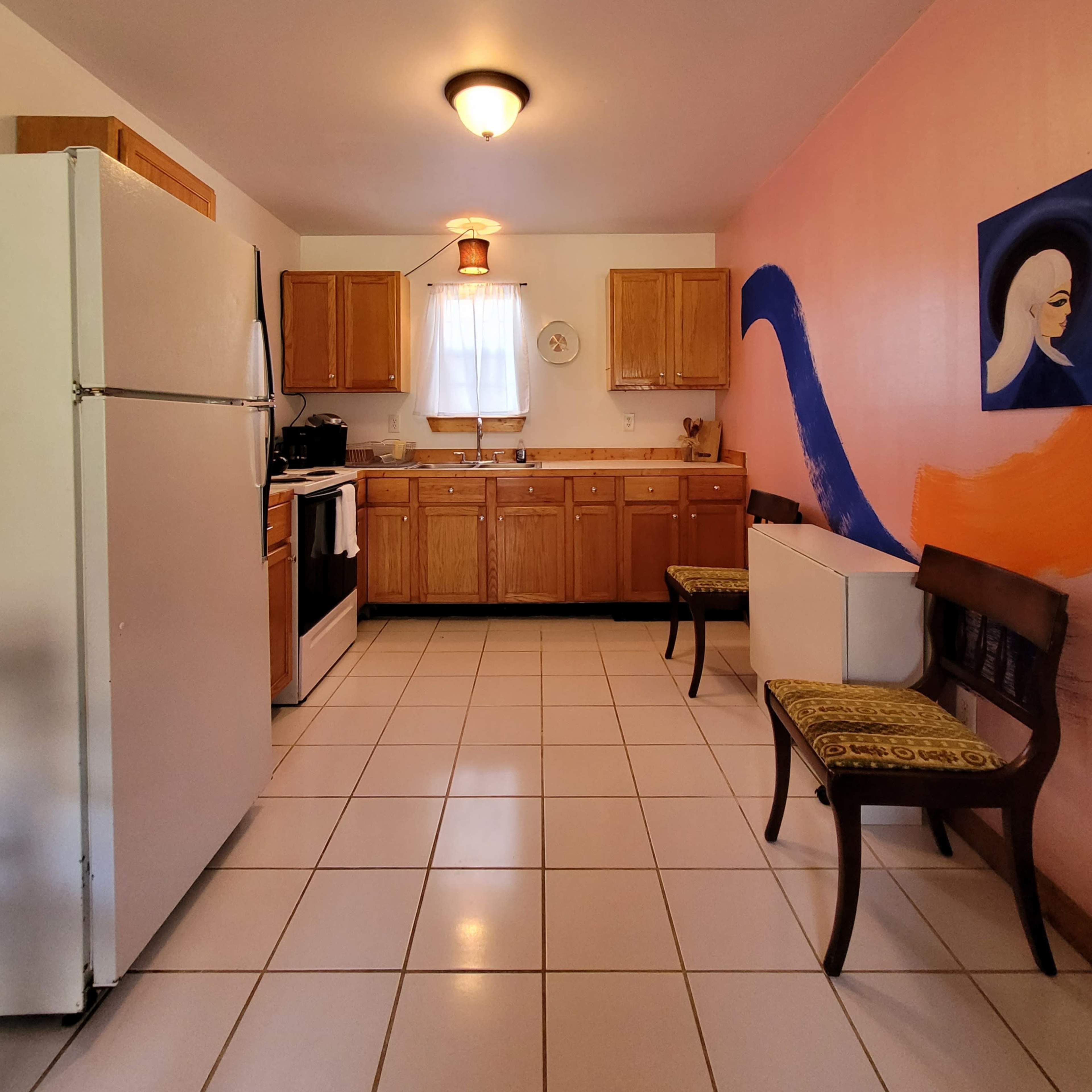 The kitchen features wooden cabinets, a white refrigerator, and two chairs against a pink wall decorated with abstract art.
