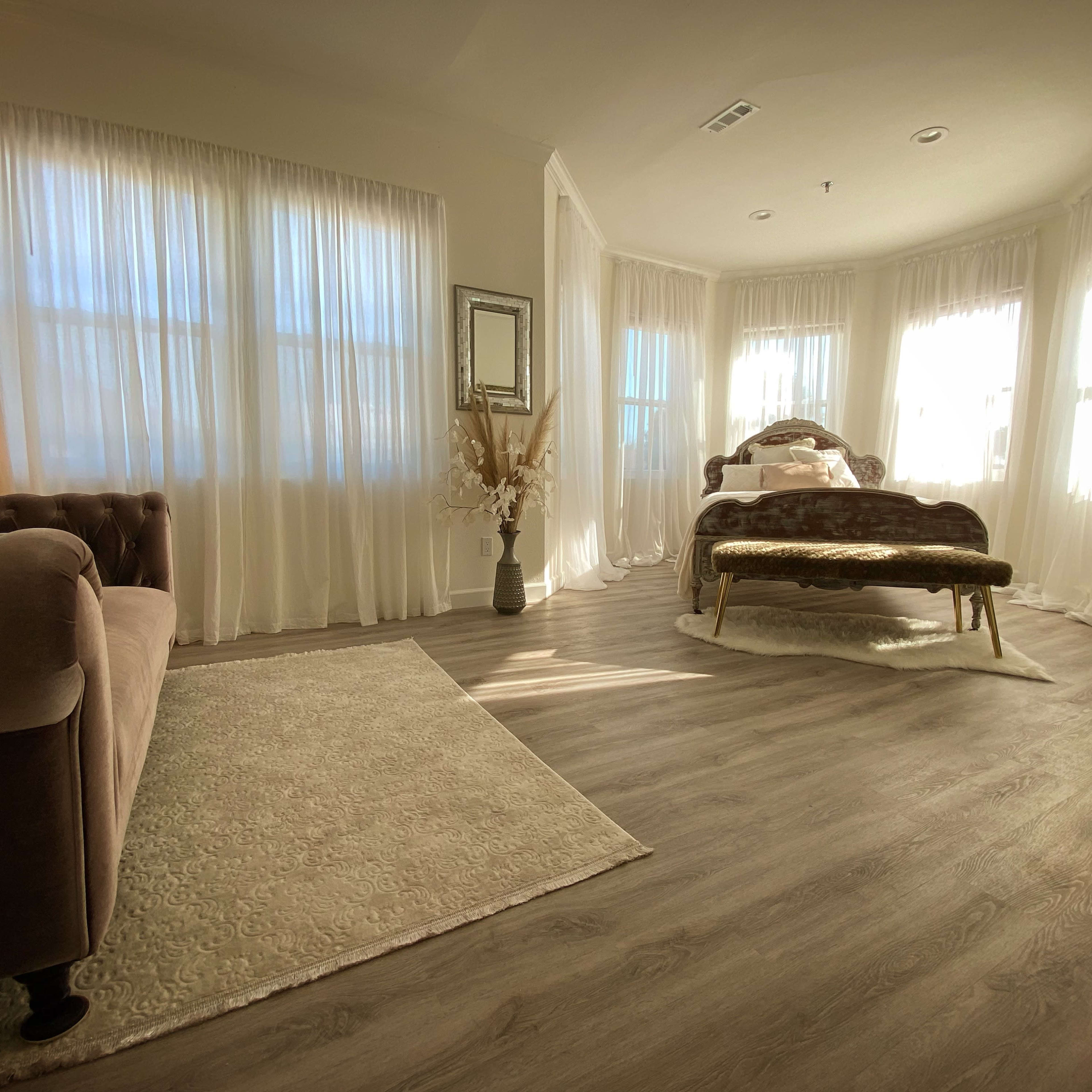 The room features a bed with an ornate headboard, a plush sofa, and large windows draped with sheer curtains that allow natural light to fill the space.
