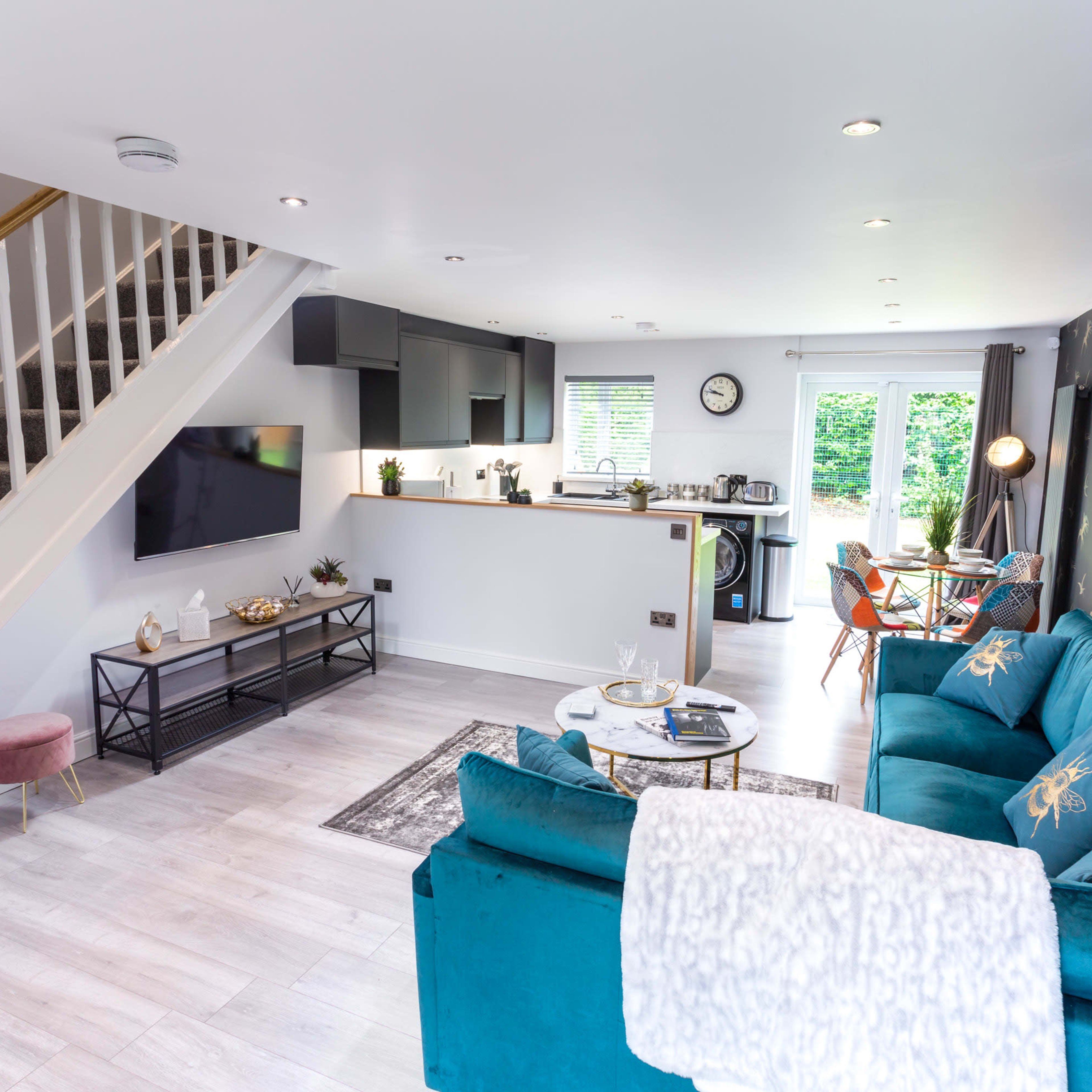 The room features an open-concept living area with a teal sofa, dining space, and a kitchen visible in the background.