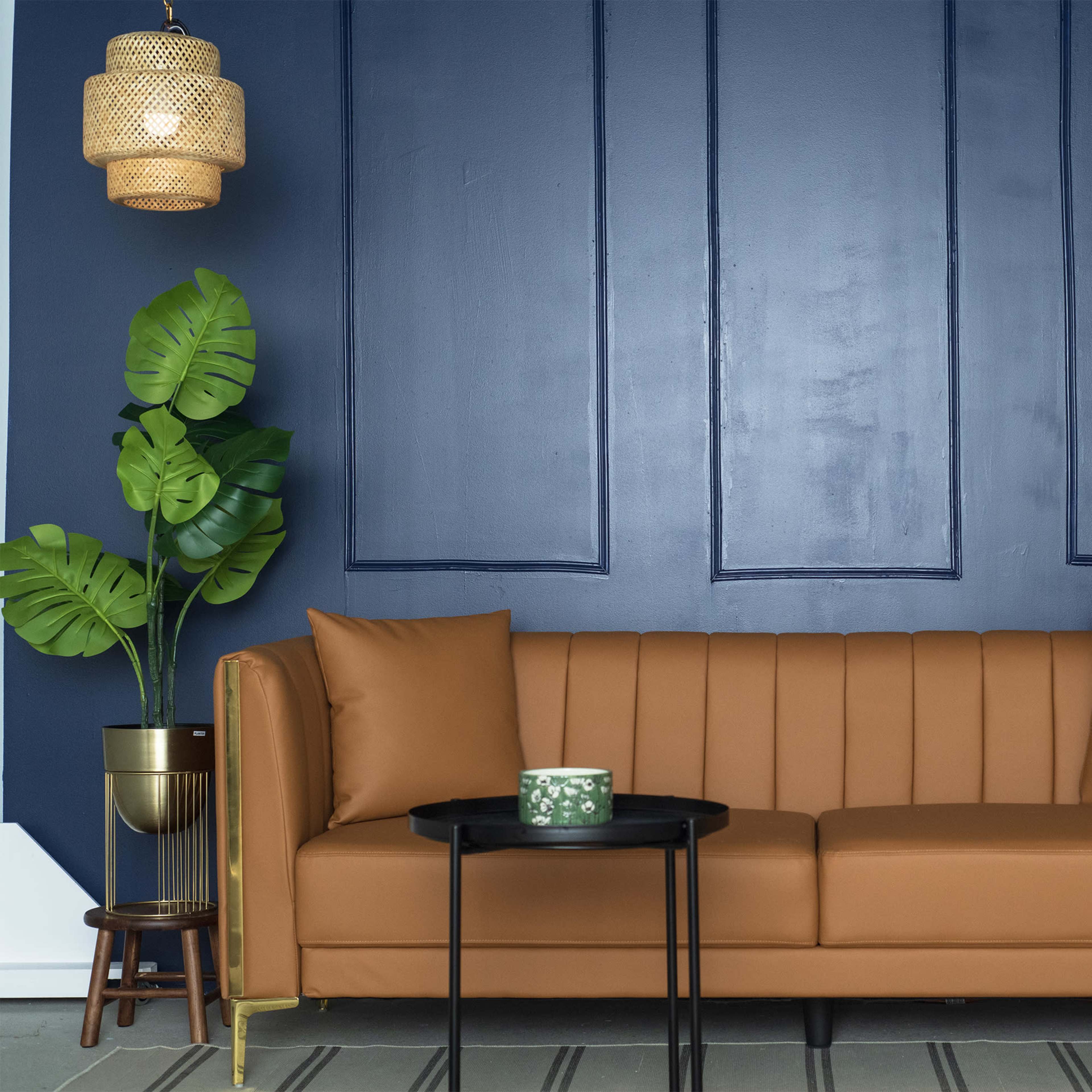 A brown upholstered sofa with two cushions is positioned against a navy blue wall, accompanied by a small black side table and a potted plant.