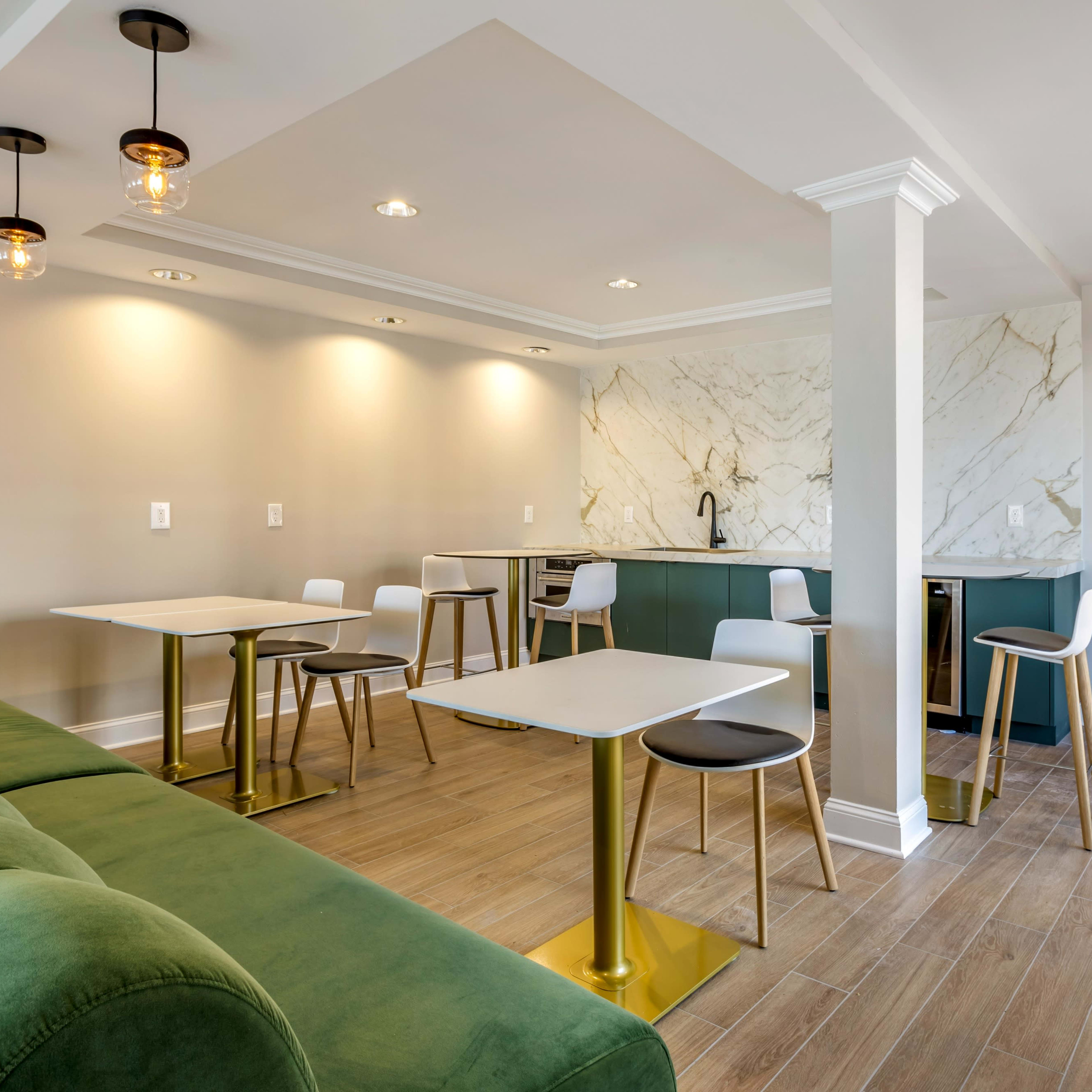 A modern, well-lit lounge area featuring green sofas, sleek tables with gold legs, and a kitchen counter with marble accents.