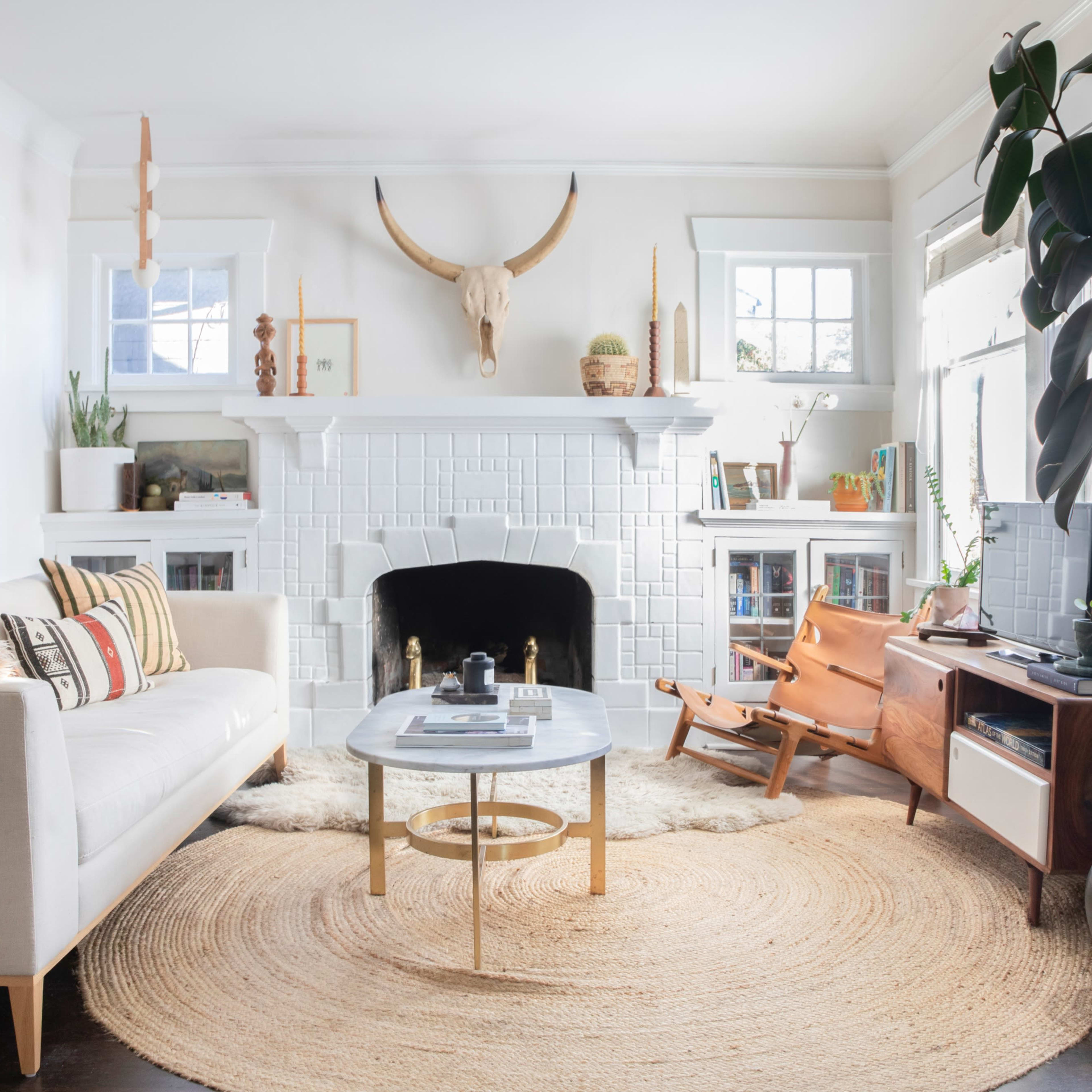 A bright living room features a white sofa, a round coffee table, a tan rug, and a fireplace adorned with decorative items and a mounted animal skull.