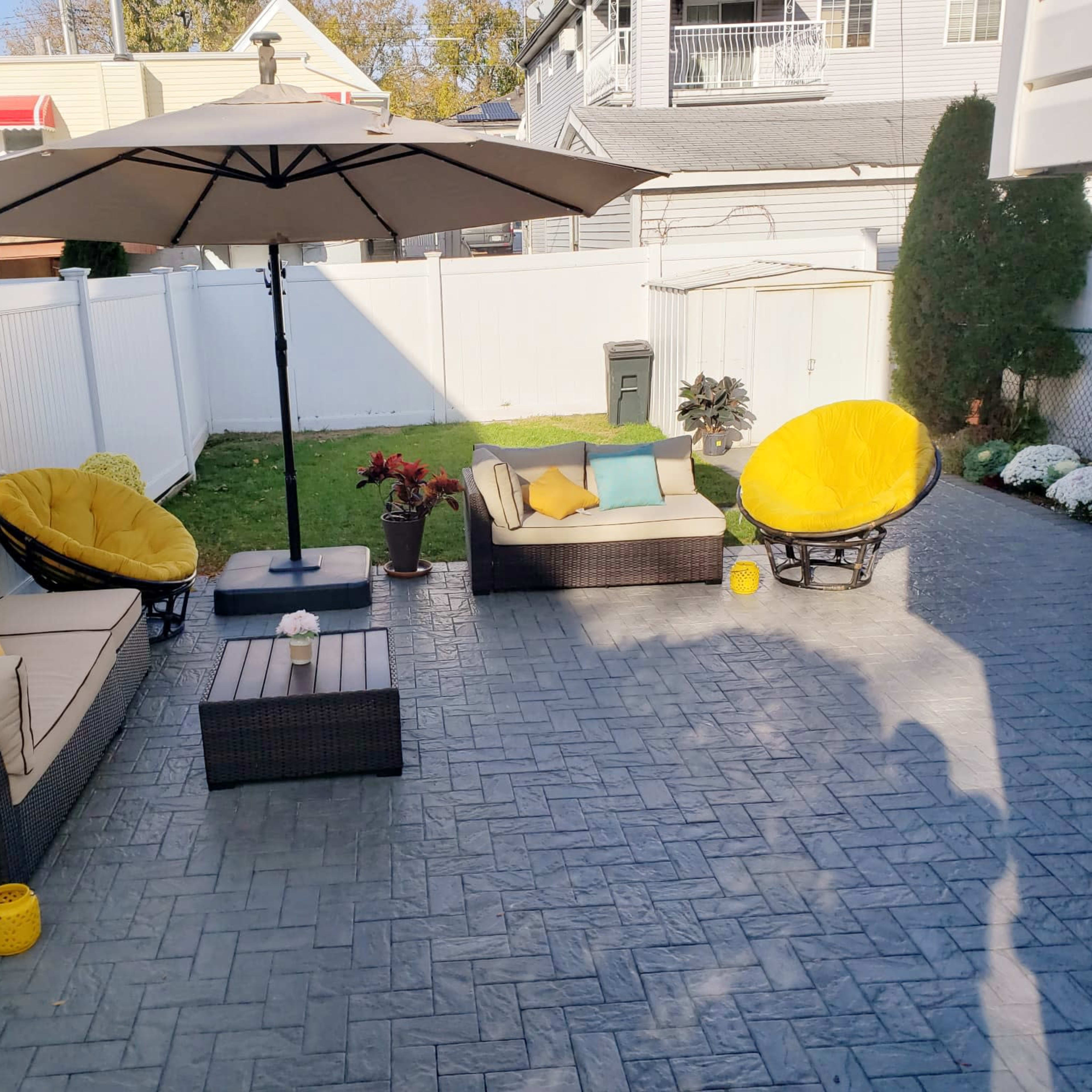 A backyard patio with two yellow cushioned lounge chairs, two beige sofas, a central coffee table, and a large umbrella, surrounded by a white fence and greenery.