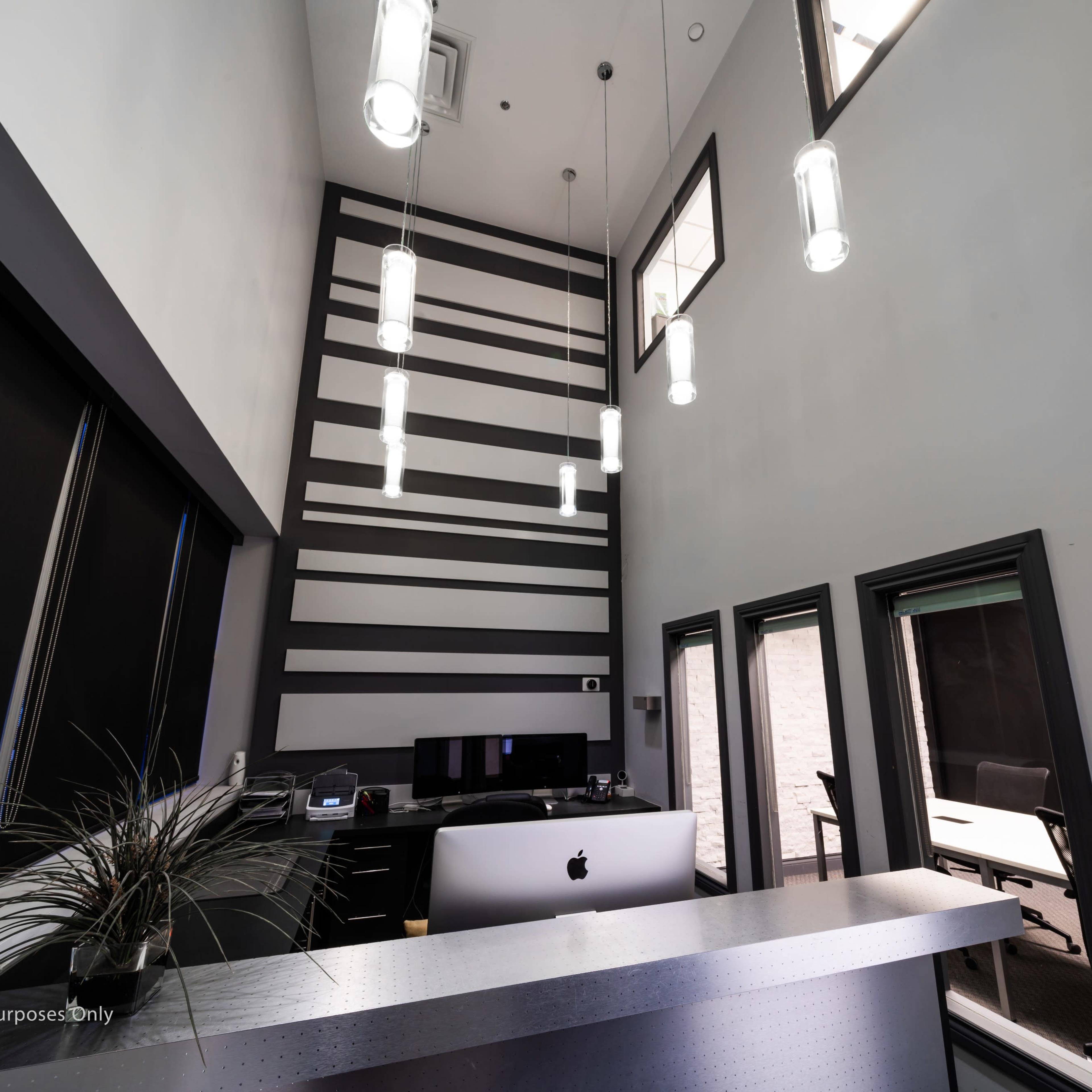 A modern office interior featuring a reception desk with an Apple computer, high ceilings adorned with pendant lights, and large windows on the wall.