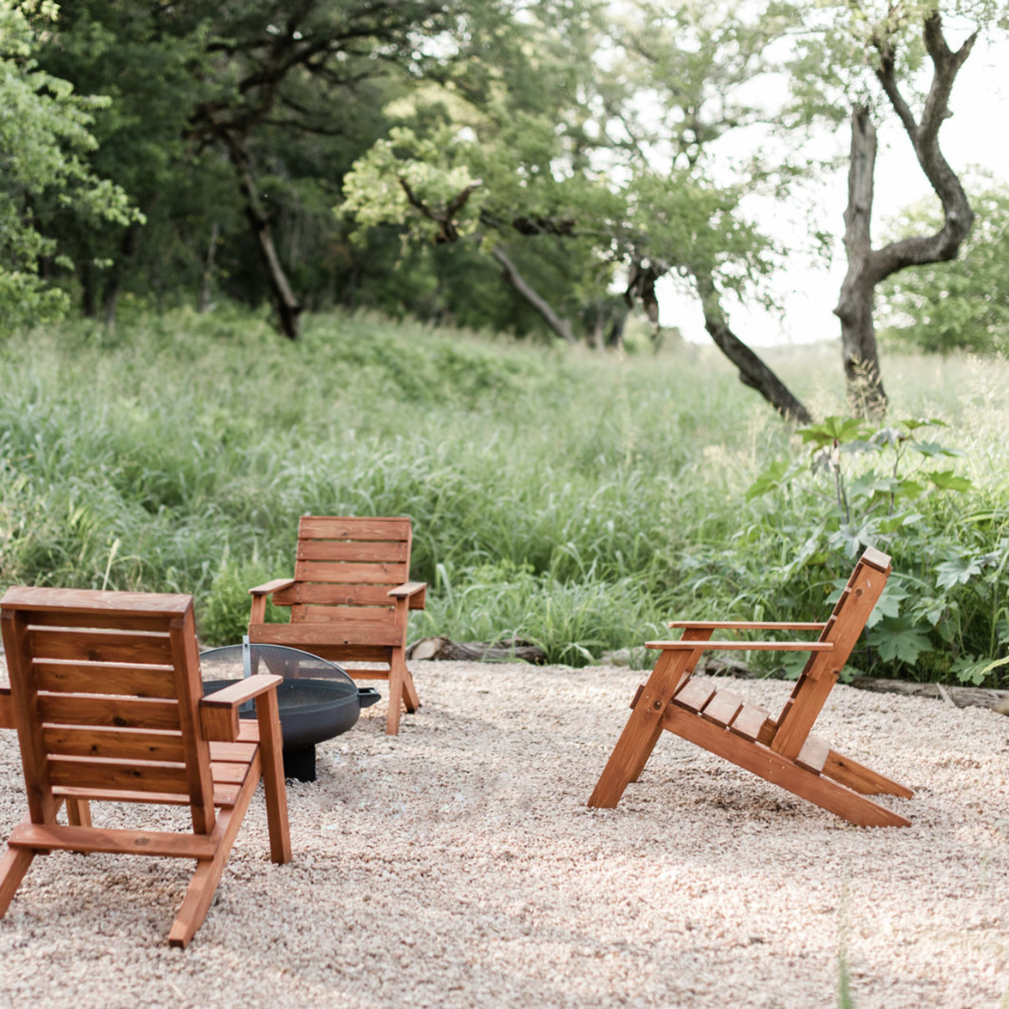 Four wooden chairs are arranged in a circle around a small fire pit on a gravel area surrounded by tall grass and trees.