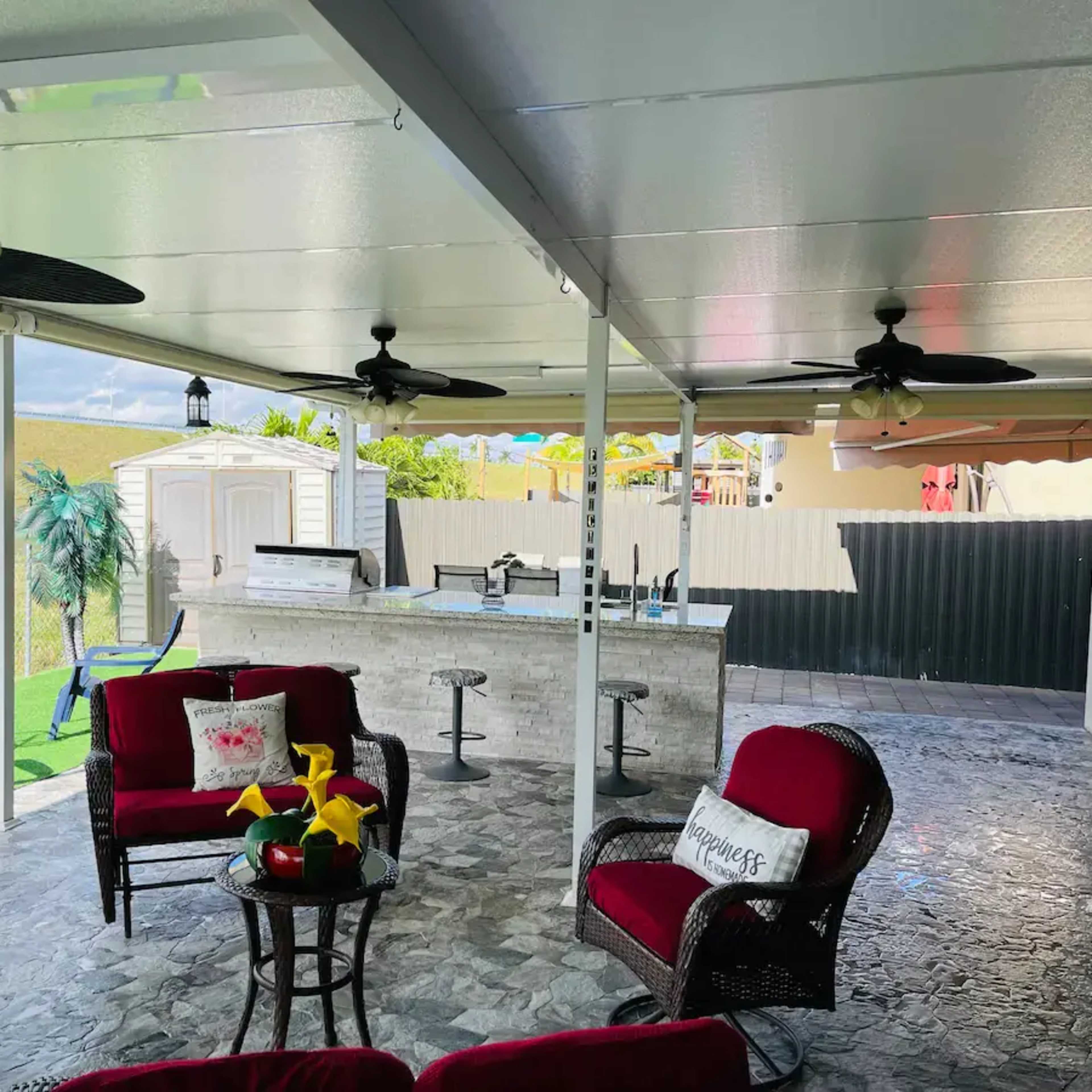 A covered outdoor patio with red seating, a small table, a bar area, and a well-maintained lawn.