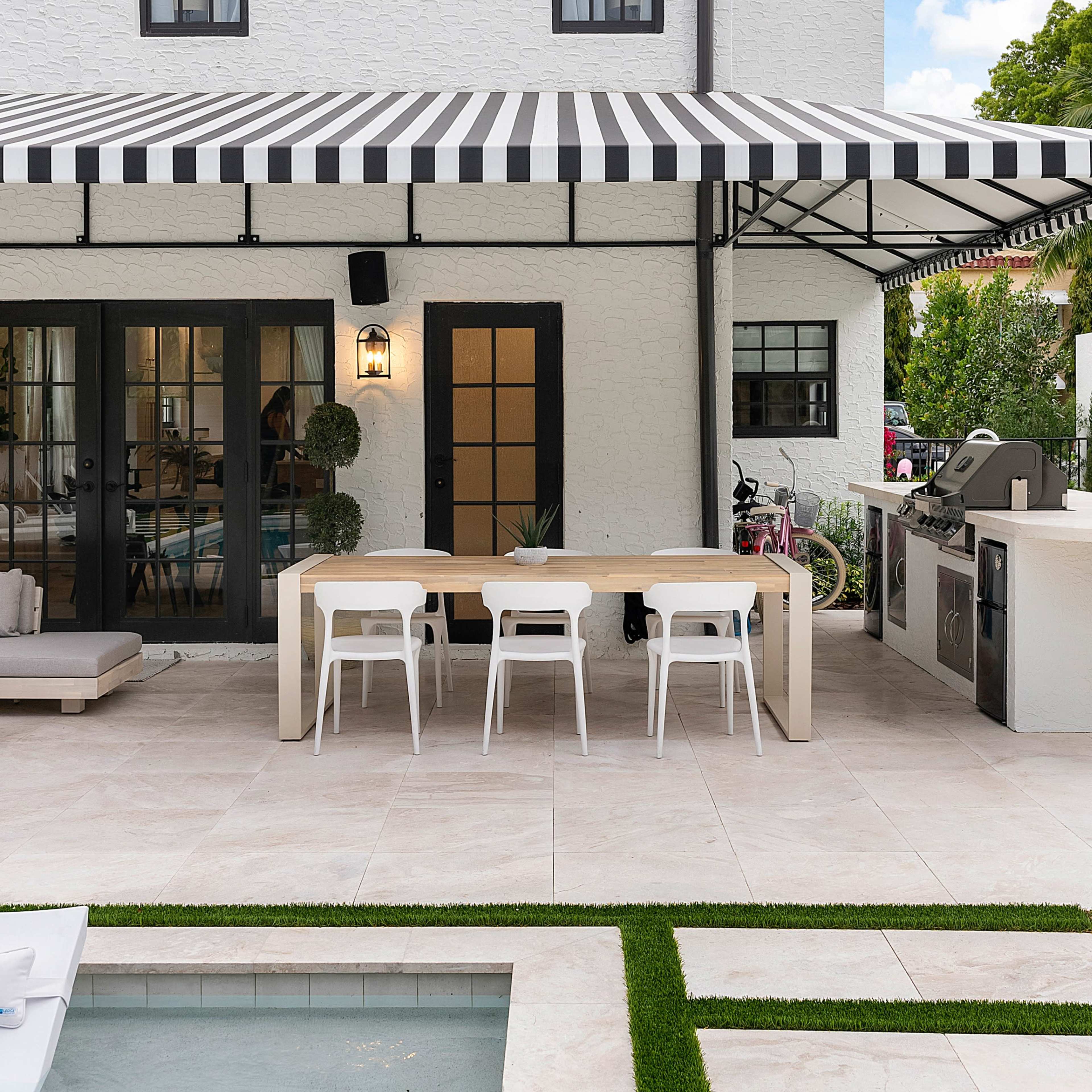 A modern outdoor dining area with a striped awning, a rectangular table, and white chairs beside a swimming pool and landscaped grass.